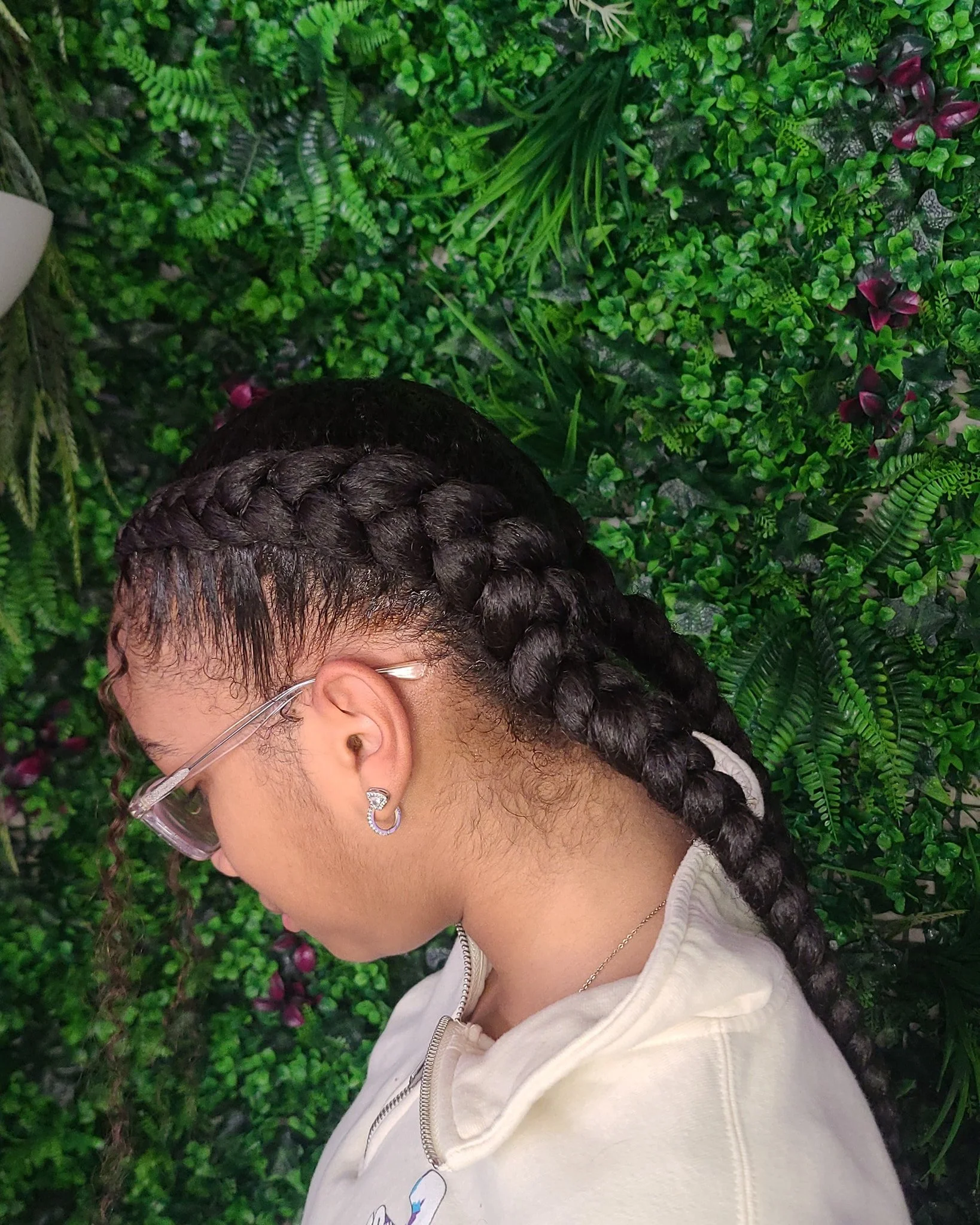 Young woman with long braided hair, silver earrings, glasses, and a white hoodie, standing against a lush green floral wall.