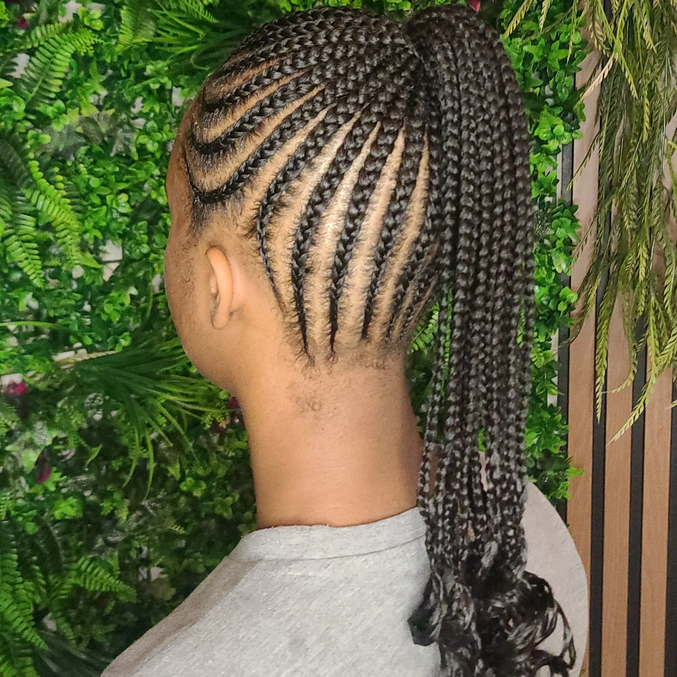 Back view of a woman with intricately styled cornrows and braided ponytail, with a backdrop of green plants.