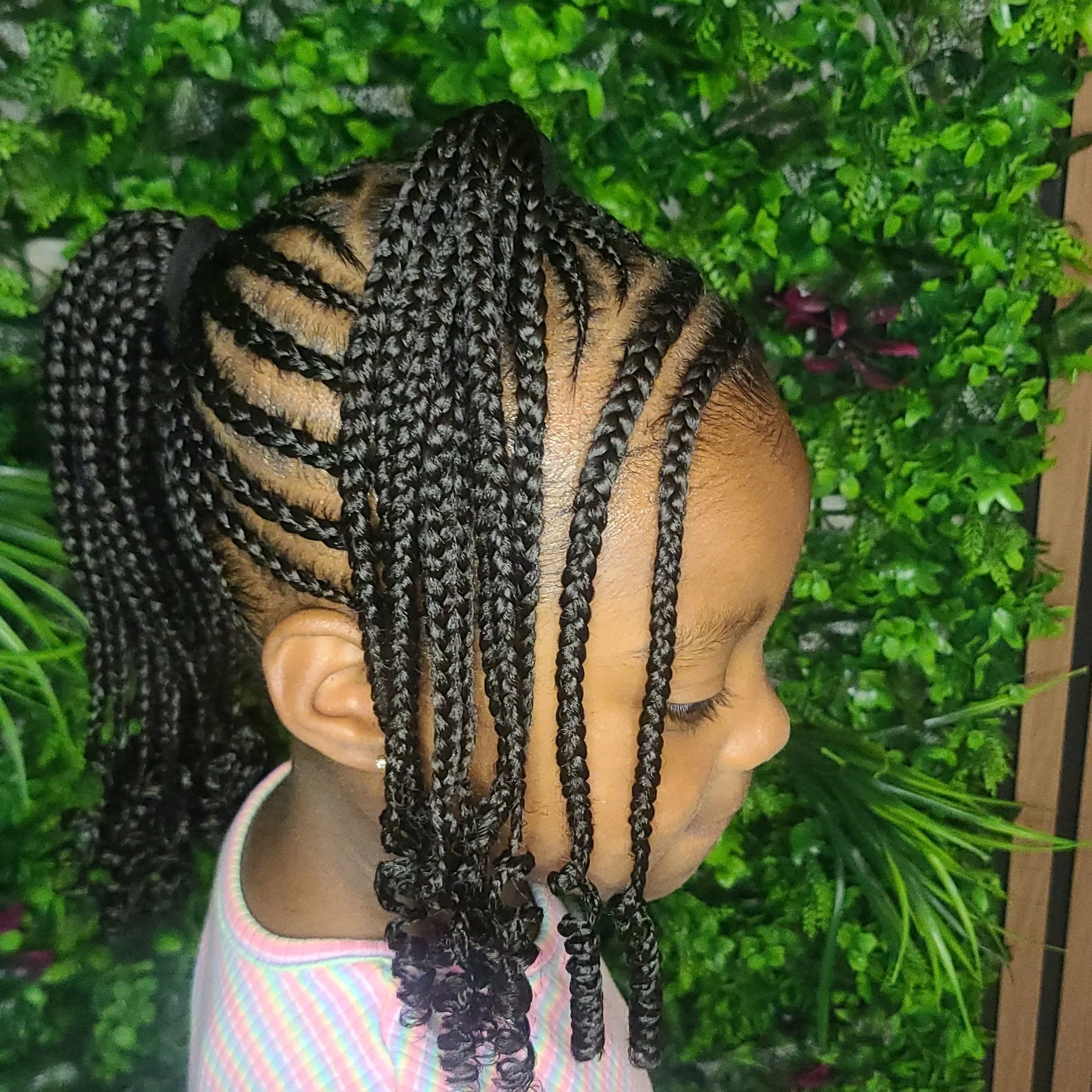 Side profile of a young girl with braided hair, wearing a colorful striped shirt, standing against a green leafy background.