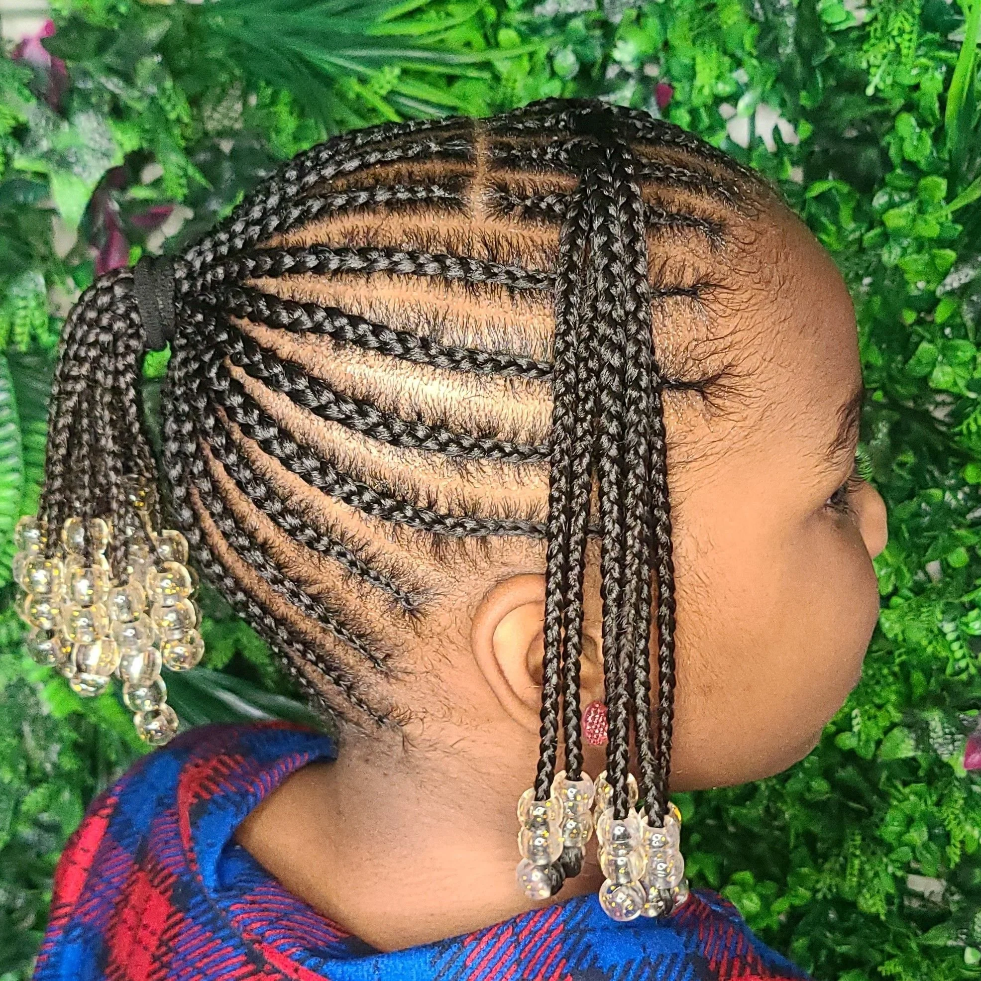 A young girl showcases a hairstyle with tightly braided cornrows decorated with clear beads and gold accents, against a background of green leaves.