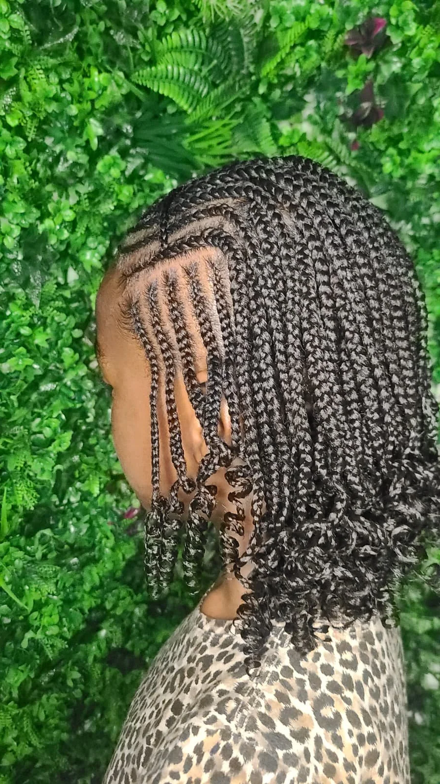 Side view of a person with tightly braided hair, wearing a leopard print top, standing in front of green foliage.