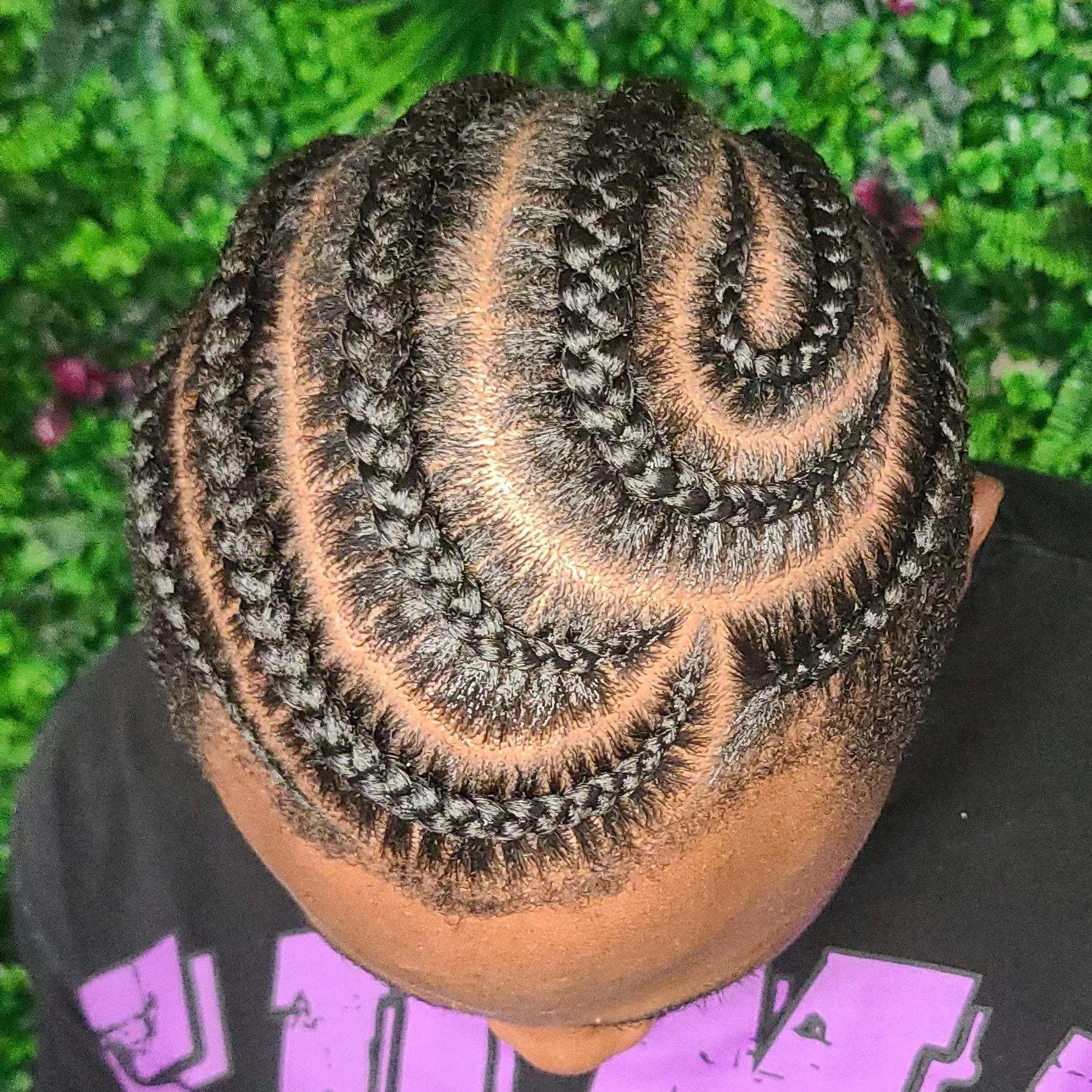 Top view of a person's head with intricate braids in a spiral pattern, featuring neat parts and shiny hair, against a background of green foliage.