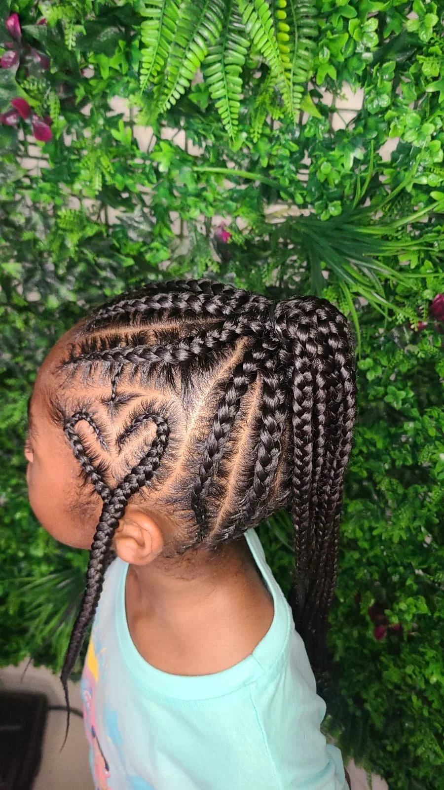 A young girl with intricate braided hairstyle adorned with a heart pattern, standing against a lush green plant backdrop.