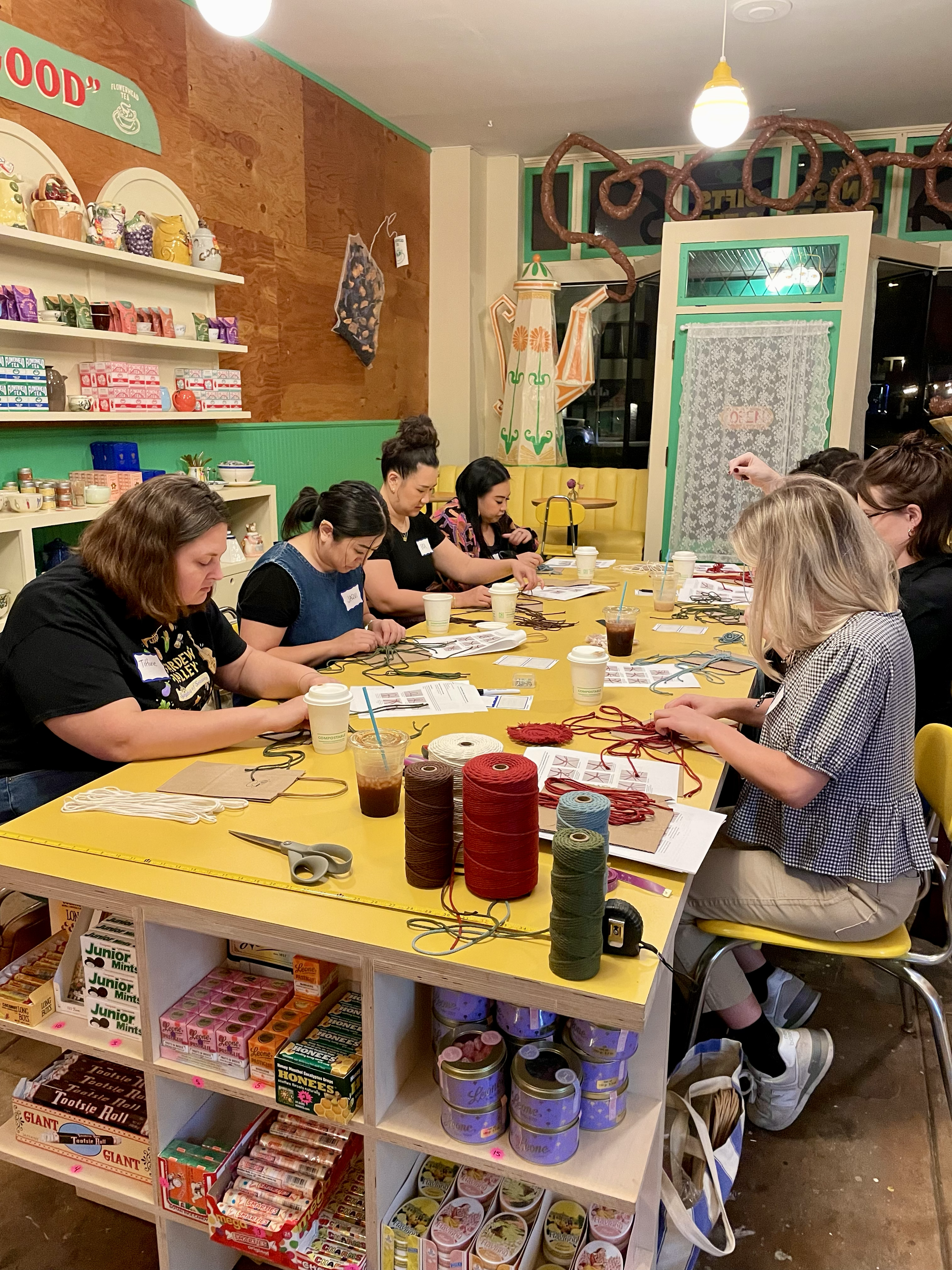 A group of women sitting around a yellow table, engaged in a crafting activity with yarns of different colors, in a cozy, decorated indoor space with store shelves and whimsical decorations.