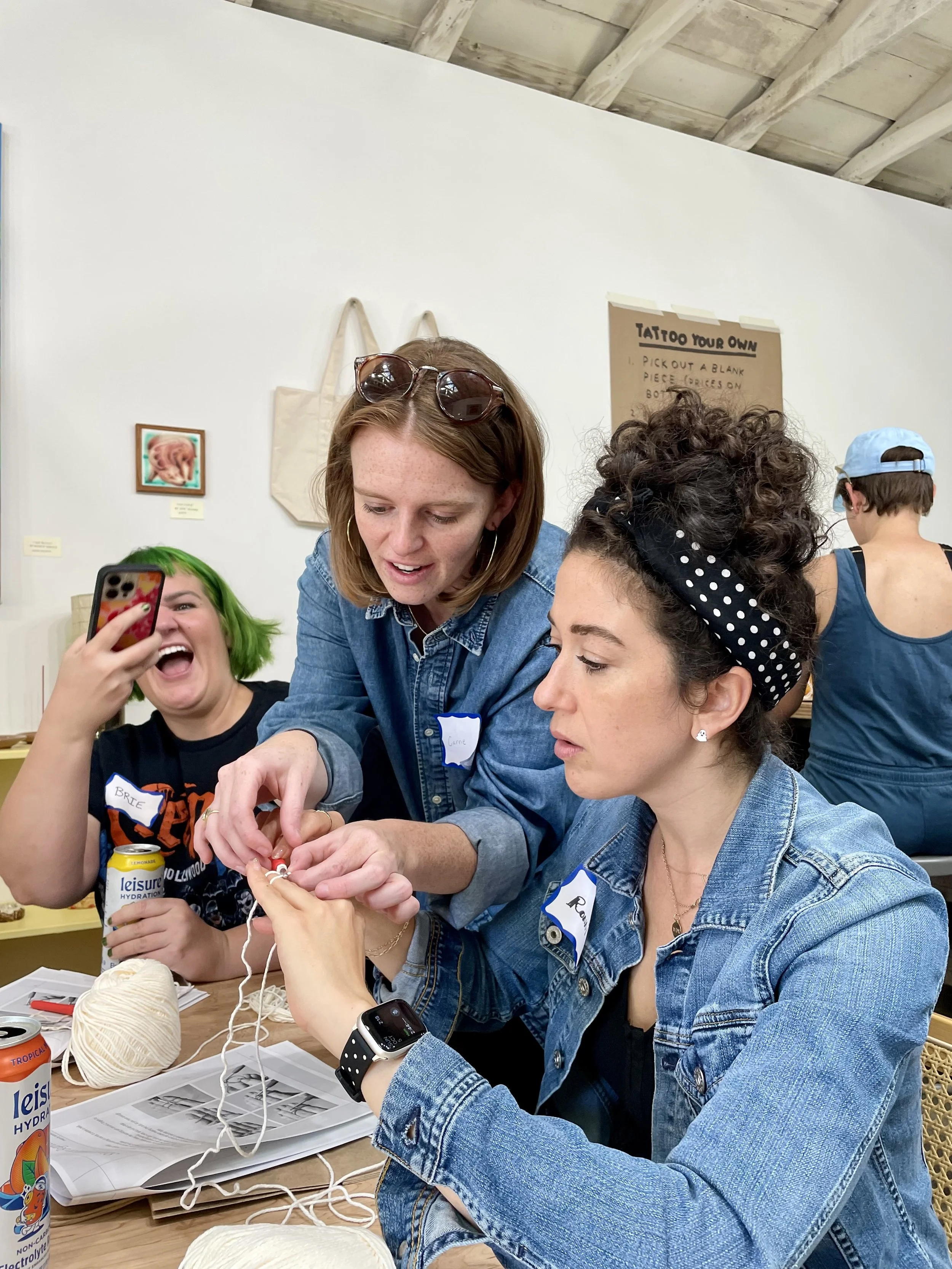 Three women are sitting at a table, one of them crocheting with yarn, one helping the crocheter, and another taking a picture of the process. They are inside a room with white walls and wooden ceiling beams.