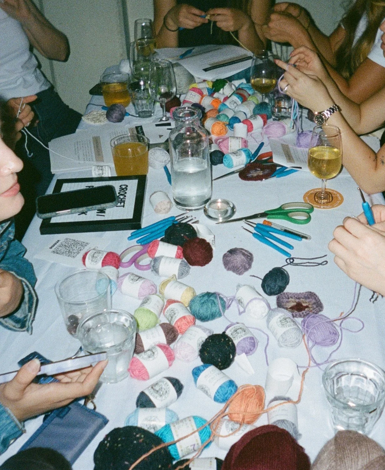 A group of people sitting around a table with various yarn balls, knitting needles, scissors, glasses of drinks, and papers, engaging in a knitting or crafting activity.