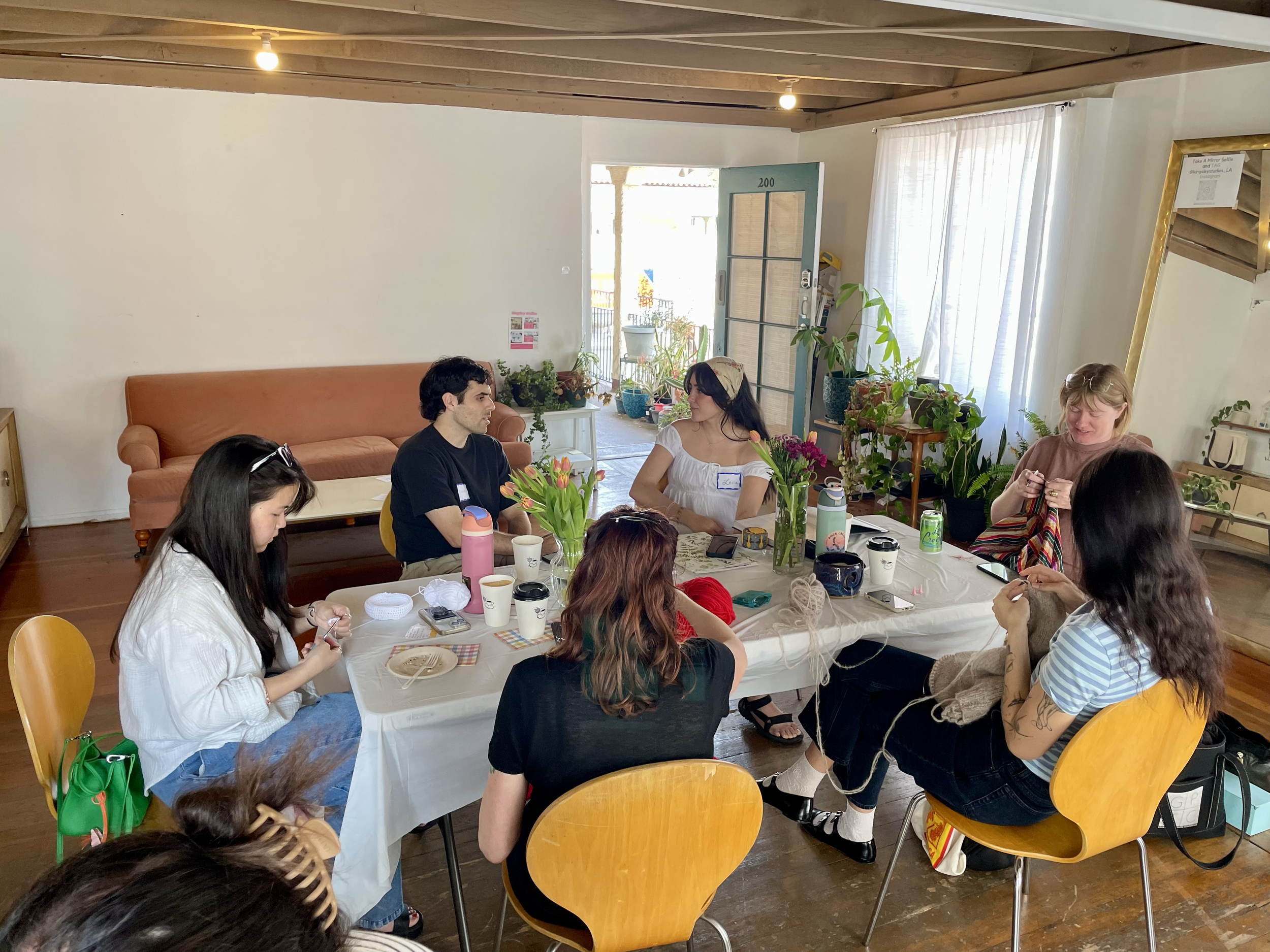 A group of women and one man sit around a table engaged in knitting and chatting. The table has flowers, drinks, and knitting supplies. The setting appears to be a cozy room with natural light, a wooden ceiling, and houseplants.
