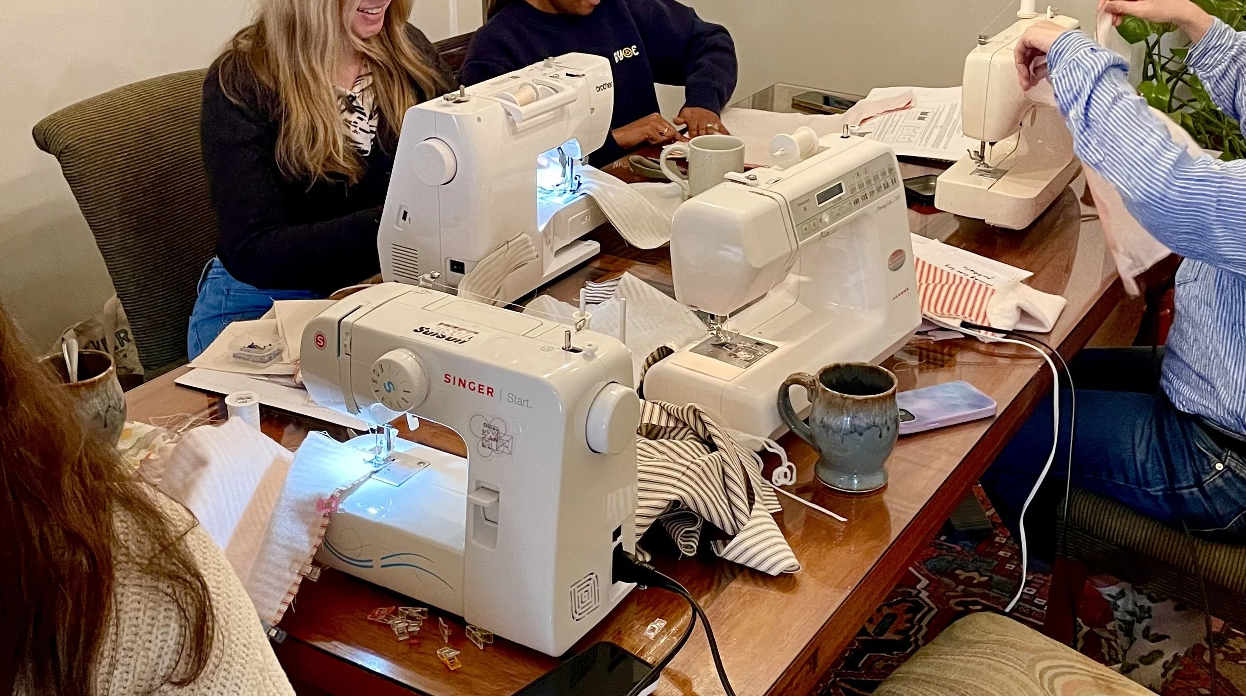 Group of people working on sewing machines at a wooden table with fabric and supplies in a cozy room.