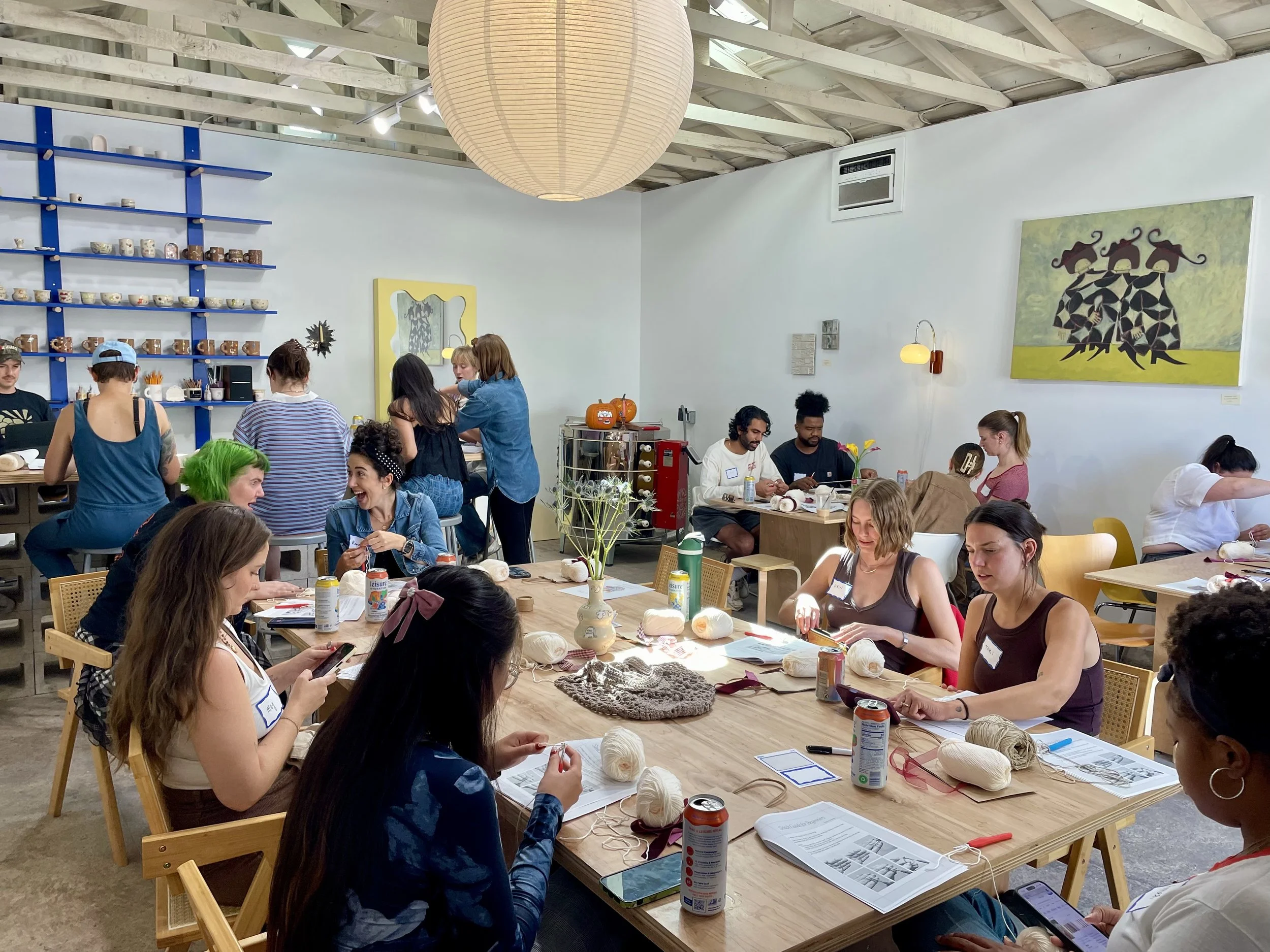 People engaging in a crafting workshop in a cozy, decorated room with shelves of cups and artwork, working with yarn and knitting supplies.