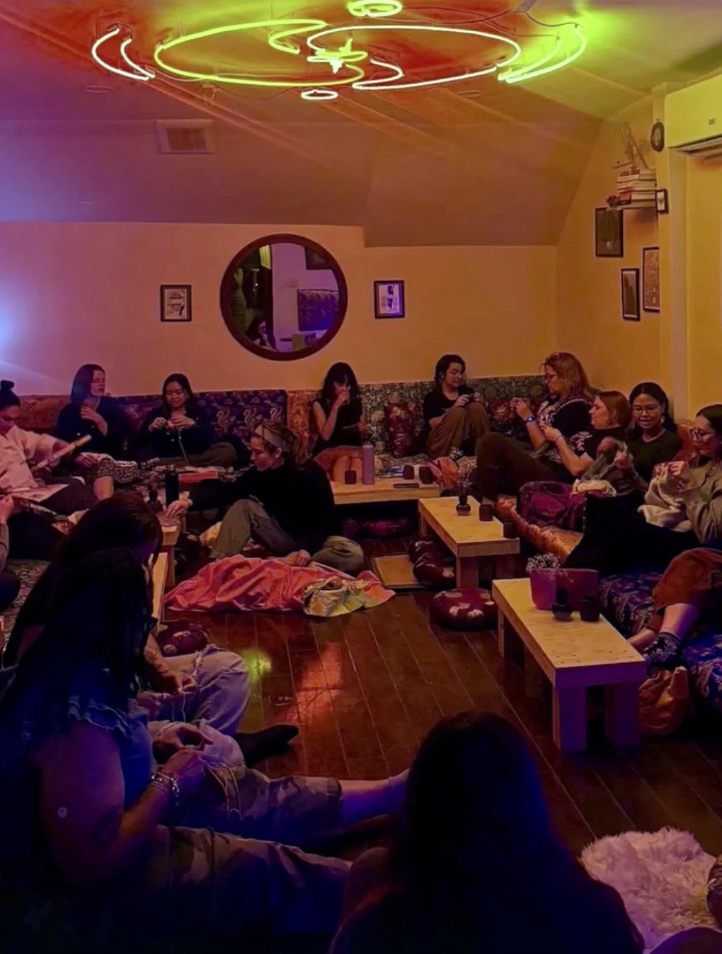 A group of people sitting on couches and floor in a cozy room with colorful lighting, watching something on a screen. The room has a round mirror on the wall, framed pictures, and wooden furniture.