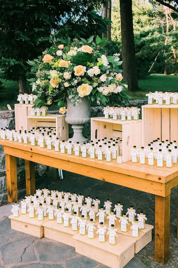 Table with small, white bottles with black caps, arranged in rows. Large floral arrangement with yellow and white flowers in a gray urn, and wooden crates on both sides of the table.