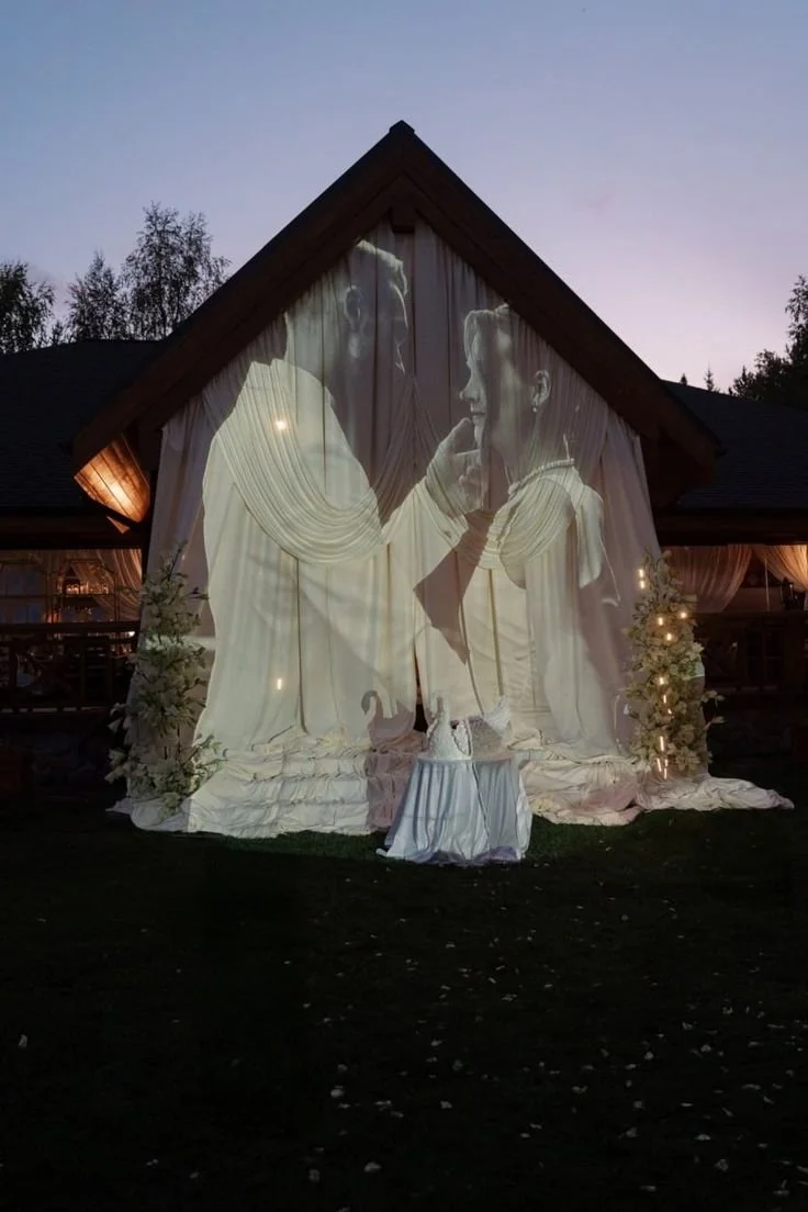 A large outdoor wedding or event setup on a house's front yard with a projection of a couple facing each other, appearing close and intimate, on a white fabric backdrop.