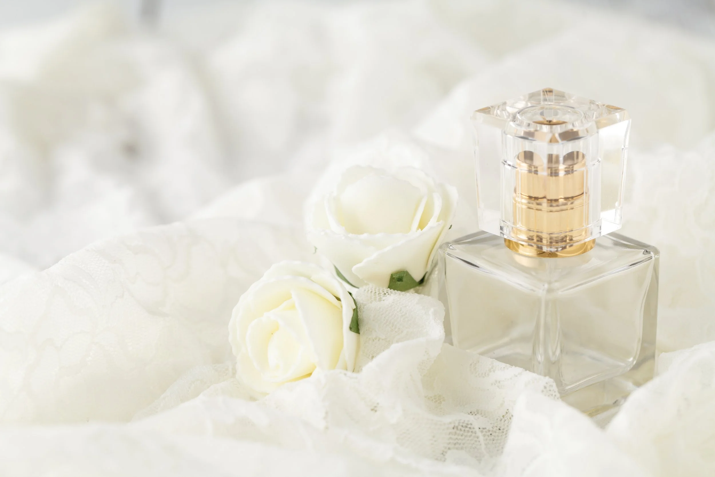 A clear glass perfume bottle with gold accents, surrounded by white roses and lace, set on a white fabric background.