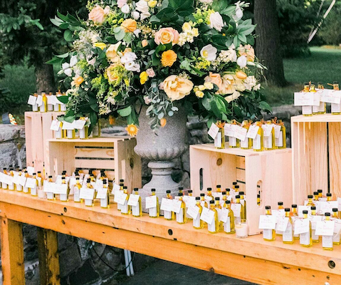 Table setup with small bottled beverages and a large floral arrangement in a gray urn at an outdoor event.