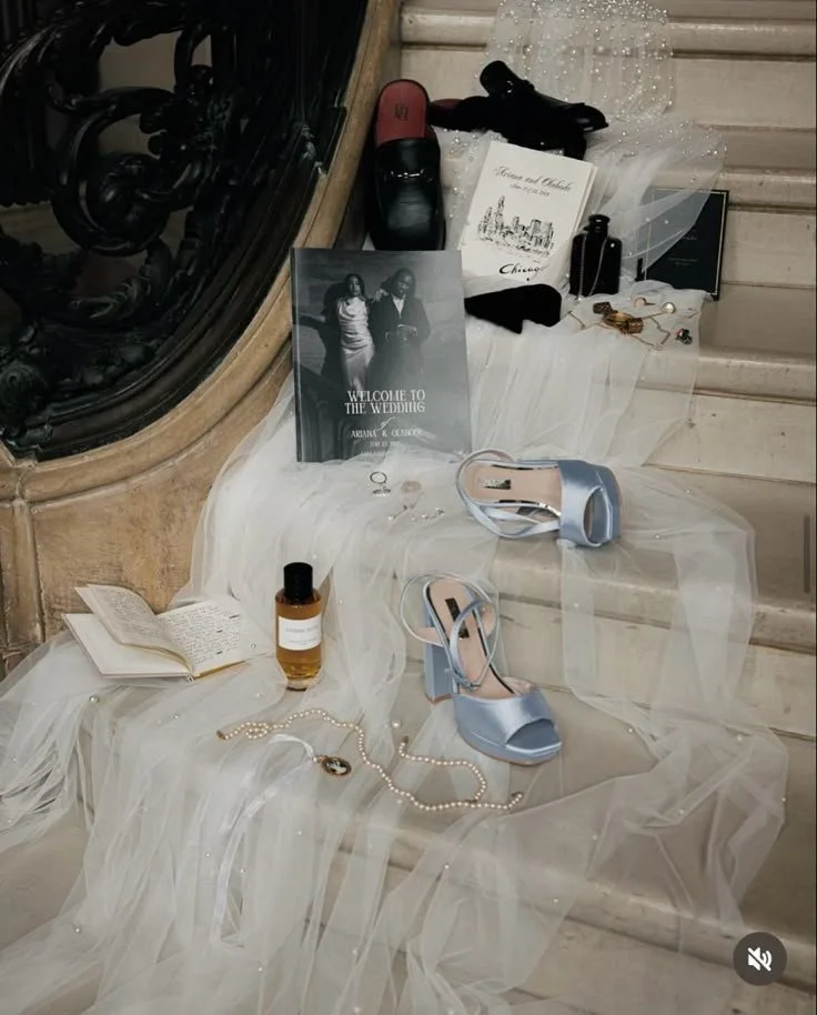 Wedding accessories and invitation displayed on a staircase, including a pair of light blue high heels, perfume, pearl necklace, black bottle, hairbrush, and a wedding program.