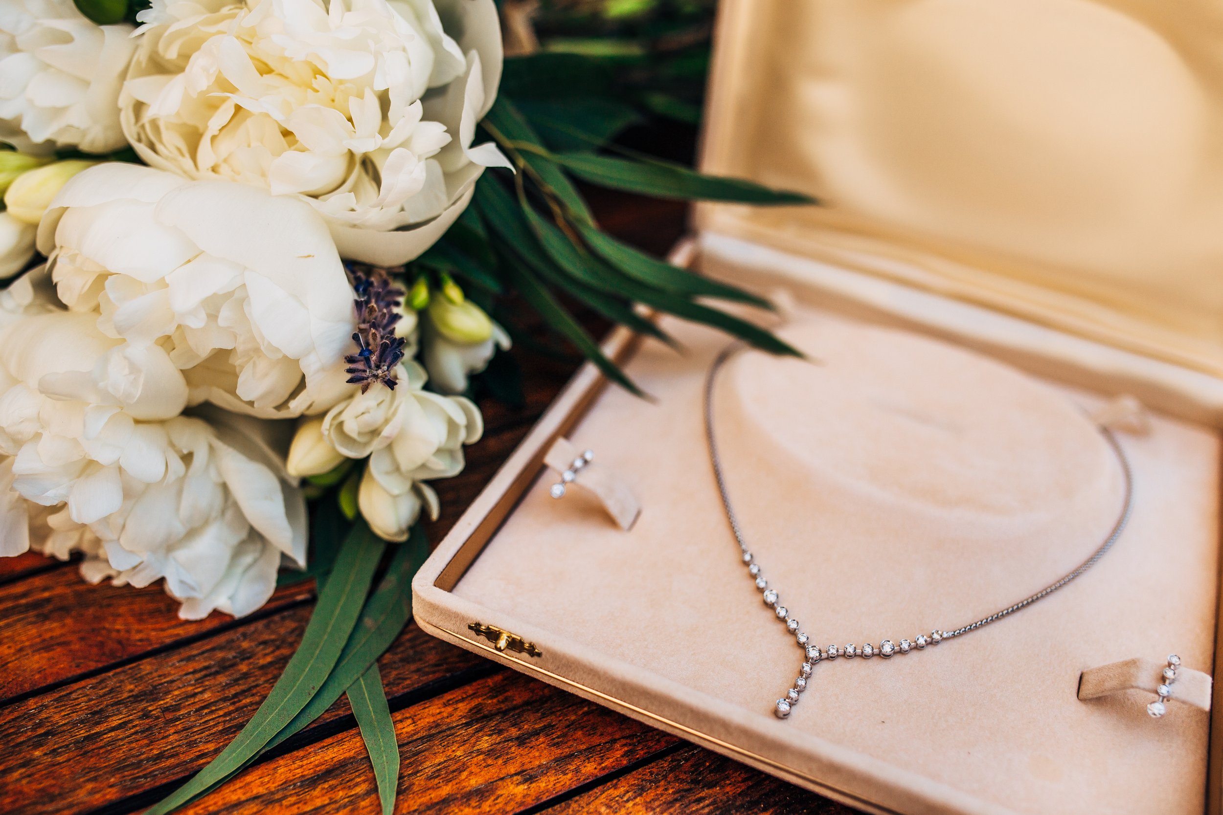 A jewelry box containing a diamond necklace and earrings placed on a wooden surface, with white flowers and green leaves in the background.