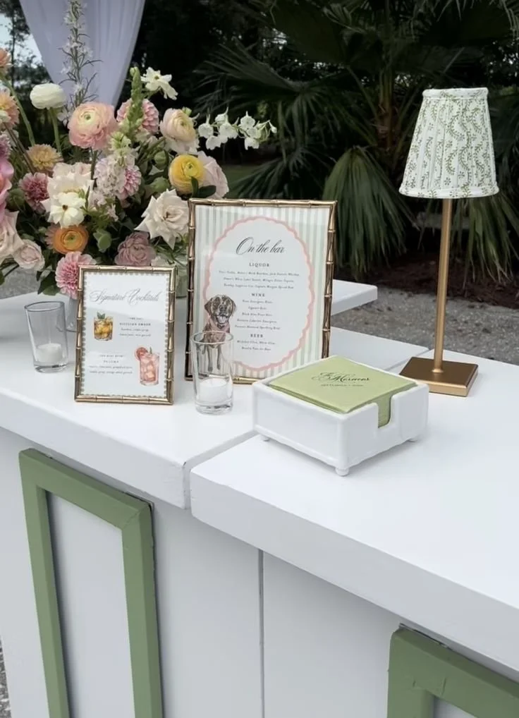 Elegant outdoor bar setup with floral arrangements, framed menus, a white tissue box, a table lamp, and small glasses on a white surface surrounded by greenery.