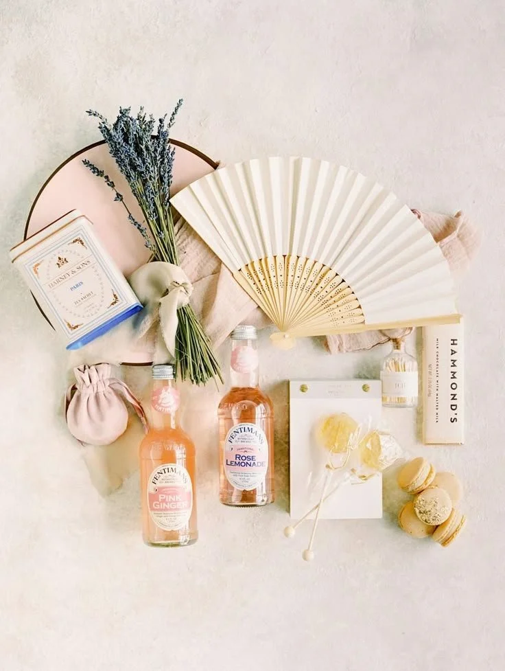 Flat lay of vintage style items including a white folding fan, lavender bouquet, pink and orange tonic water bottles, macarons, lemon candies, a pink pouch, a small gift box, and a box of tea, on a soft fabric surface.