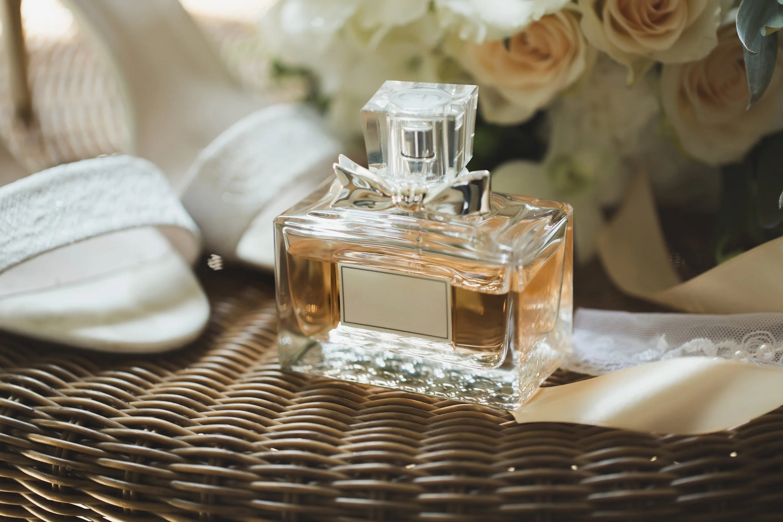 A glass perfume bottle with a bow, placed on a woven surface, surrounded by soft-focus white roses and textured fabric.