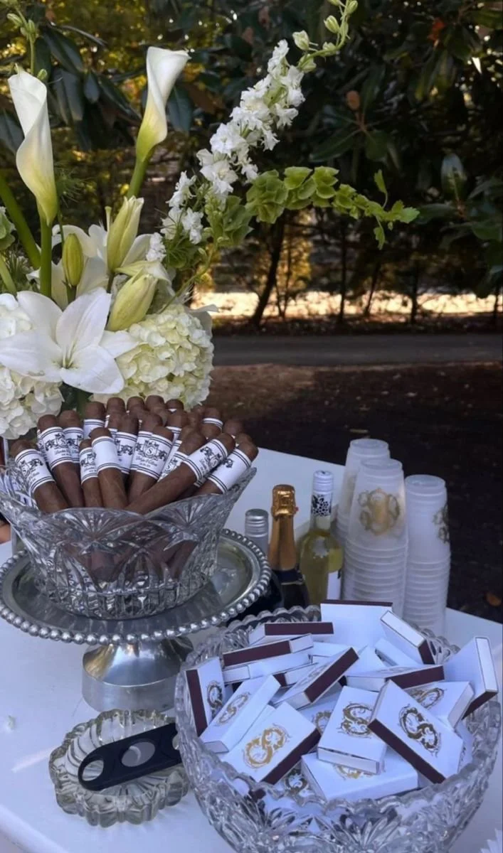 Table setup with cigars, chocolates, and beverages, decorated with white flowers including lilies and hydrangeas, in an outdoor setting.