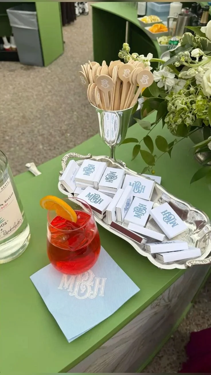 A table with a silver tray of white matchboxes, a glass with wooden stir sticks, a drink with an orange slice, and a napkin with the letters 'MH' on it, next to a floral arrangement in a vase.