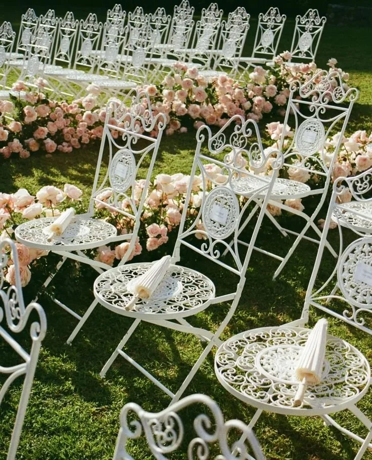White ornate metal chairs arranged for an outdoor wedding, decorated with pink flowers and small rolled white papers or programs on the seats, set on a grassy area.
