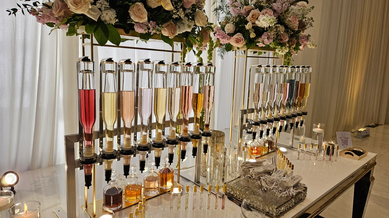 A table at an event decorated with floral arrangements on top. Multiple tall bottles contain different colored liquids and are fitted with syringes or dispensers, receiving various liquids. There are small bottles and containers, some with liquids, a