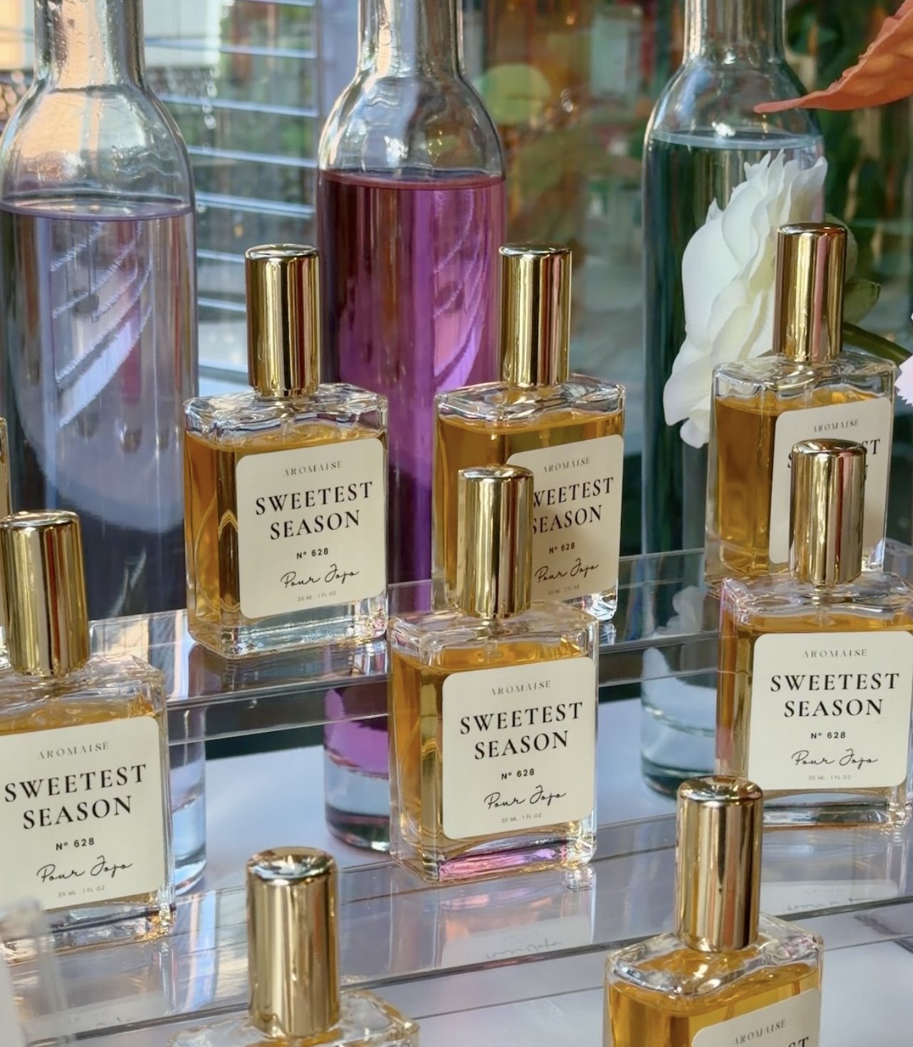 A glass perfume bottle with a bow, placed on a woven surface, surrounded by soft-focus white roses and textured fabric.