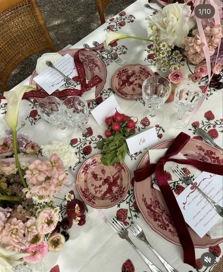 A decorated table set for a meal with pink and red themed dinnerware, floral arrangements, and place cards. It features floral-patterned plates, crystal glasses, and ribbons tied around napkins and plates.