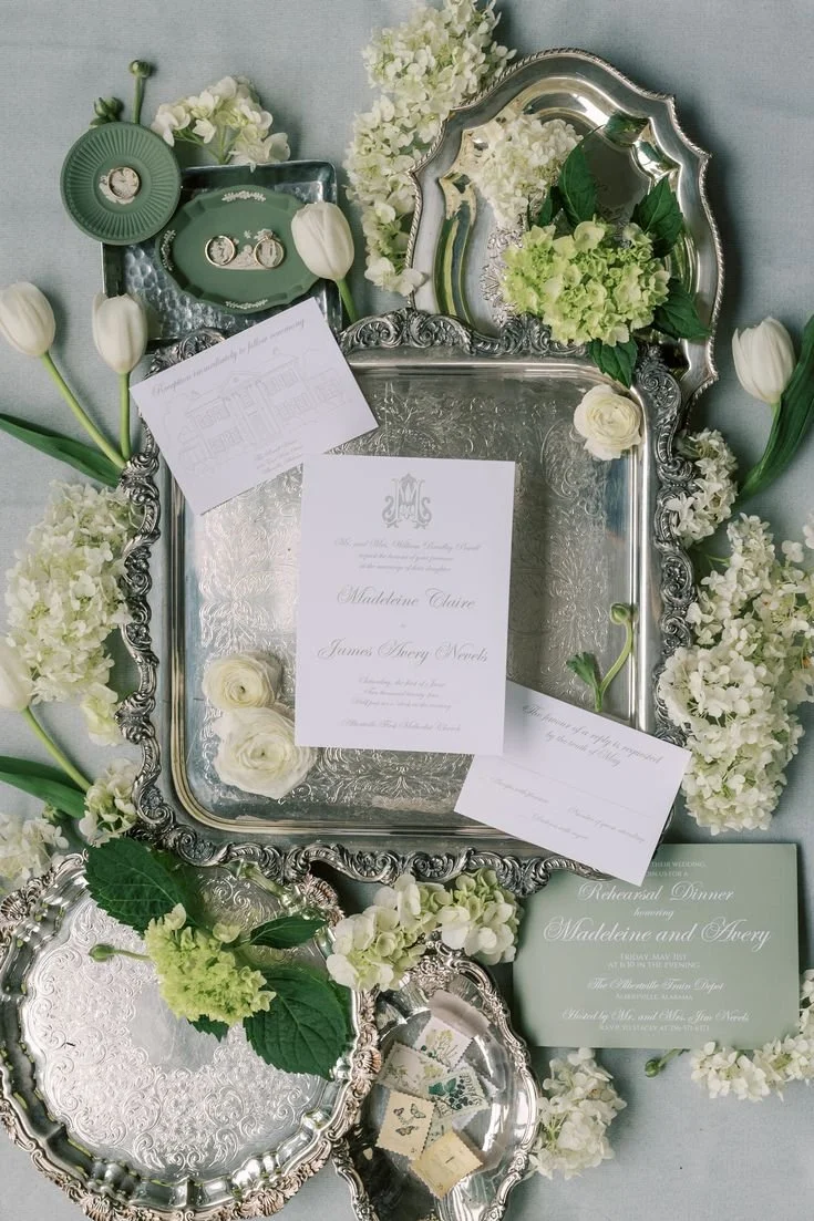 Silver serving trays and a mirror surrounded by white flowers and green leaves, with wedding invitations and notes on the trays.