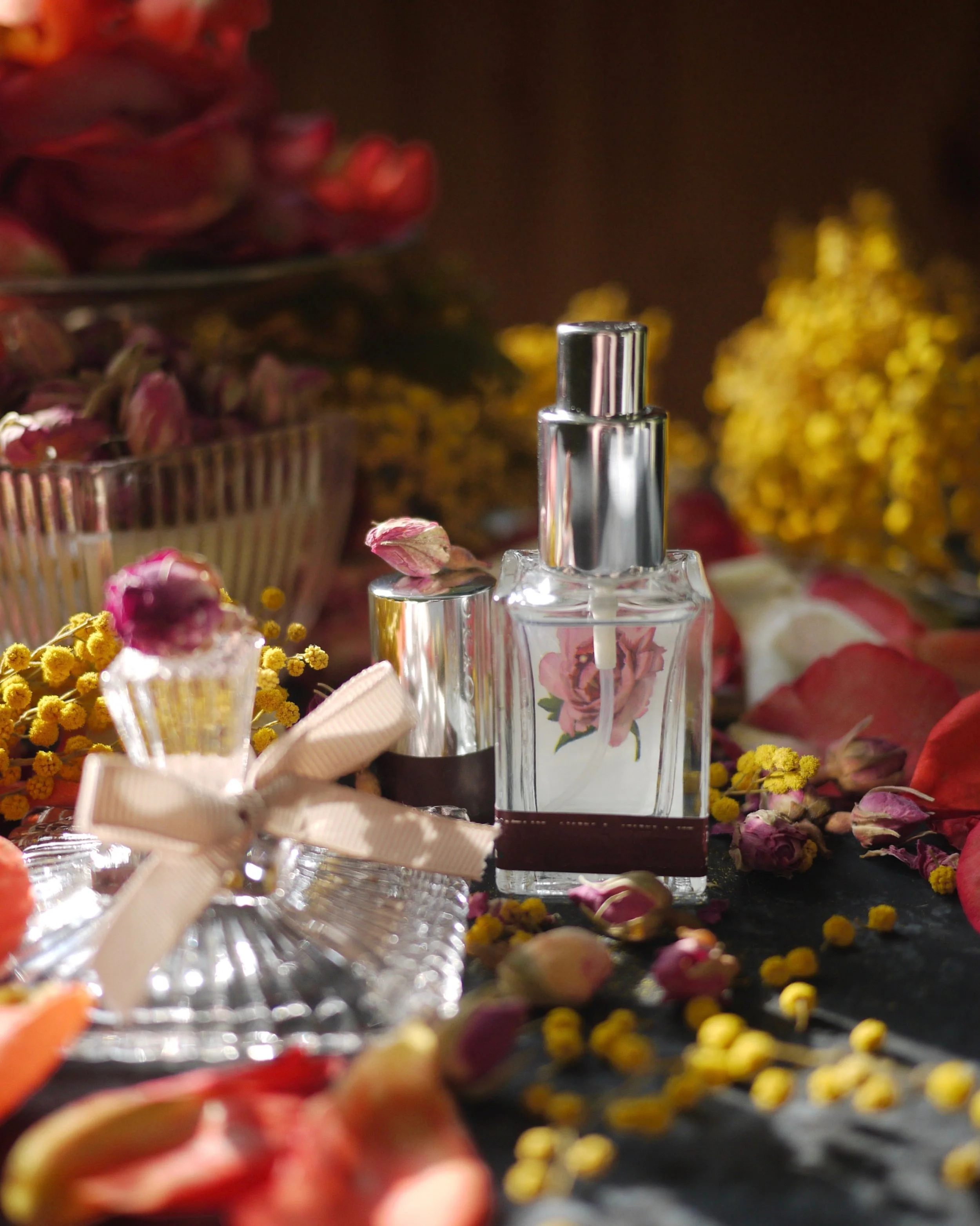 Close-up of a glass perfume bottle with a silver cap, surrounded by colorful flower petals and small yellow flowers, with additional flowers in textural baskets in the background.