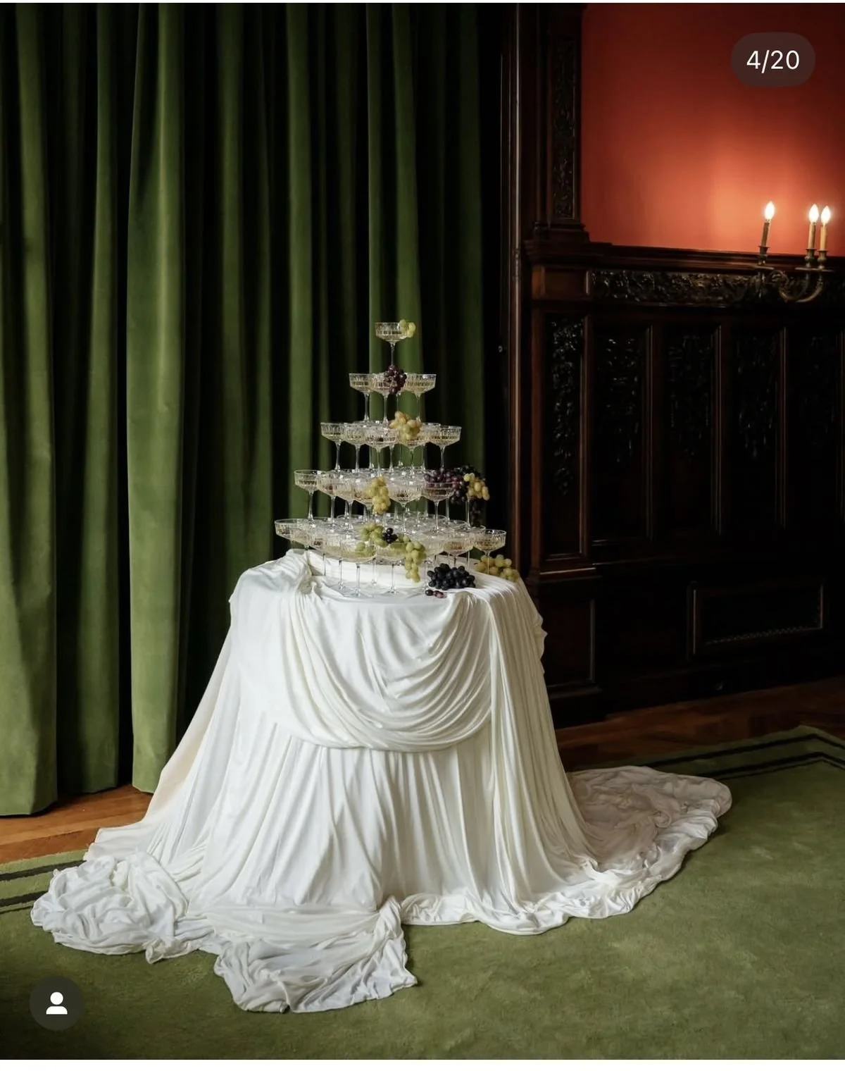 A three-tiered champagne glass tower decorated with grapes, set on a table draped with white fabric, in a room with green curtains, wooden paneling, and lit candle sconces.
