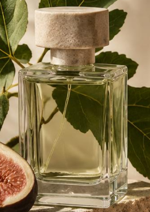 Clear glass perfume bottle with a concrete cap, surrounded by green leaves and a cut fig in the foreground.