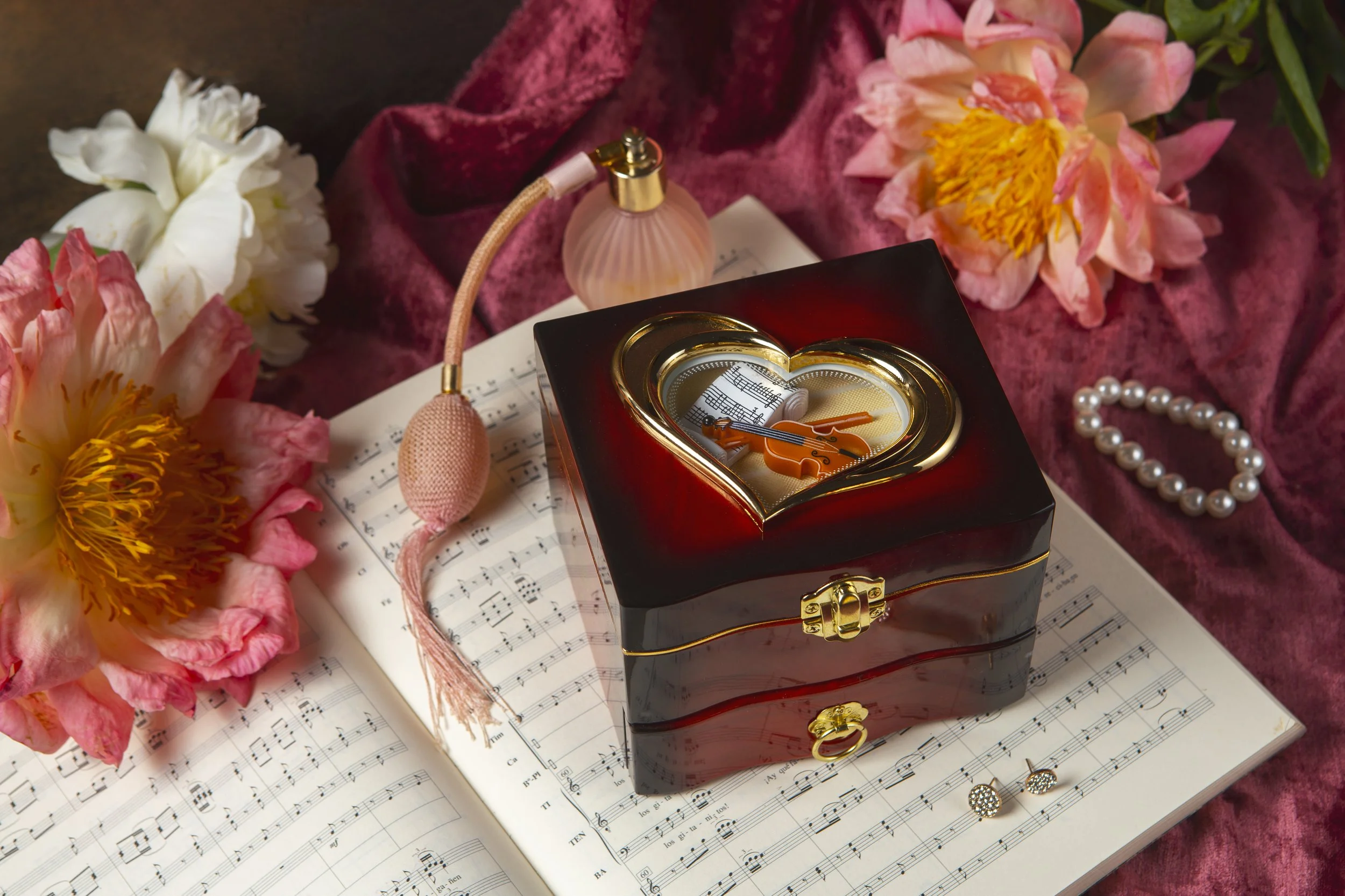 A decorative jewelry box with gold accents, shaped like a heart, is placed on an open sheet of music surrounded by pink and white peony flowers, a string of pearls, earrings, and a small pink perfume bottle.