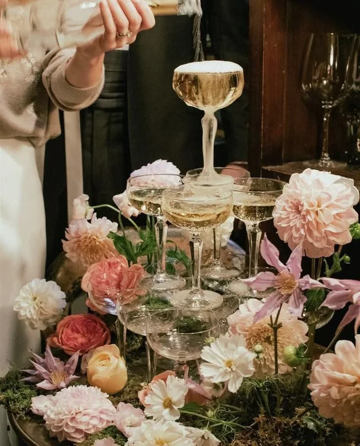 A champagne tower made of stacked glasses with floral decoration and a person pouring champagne at a celebration or special event.