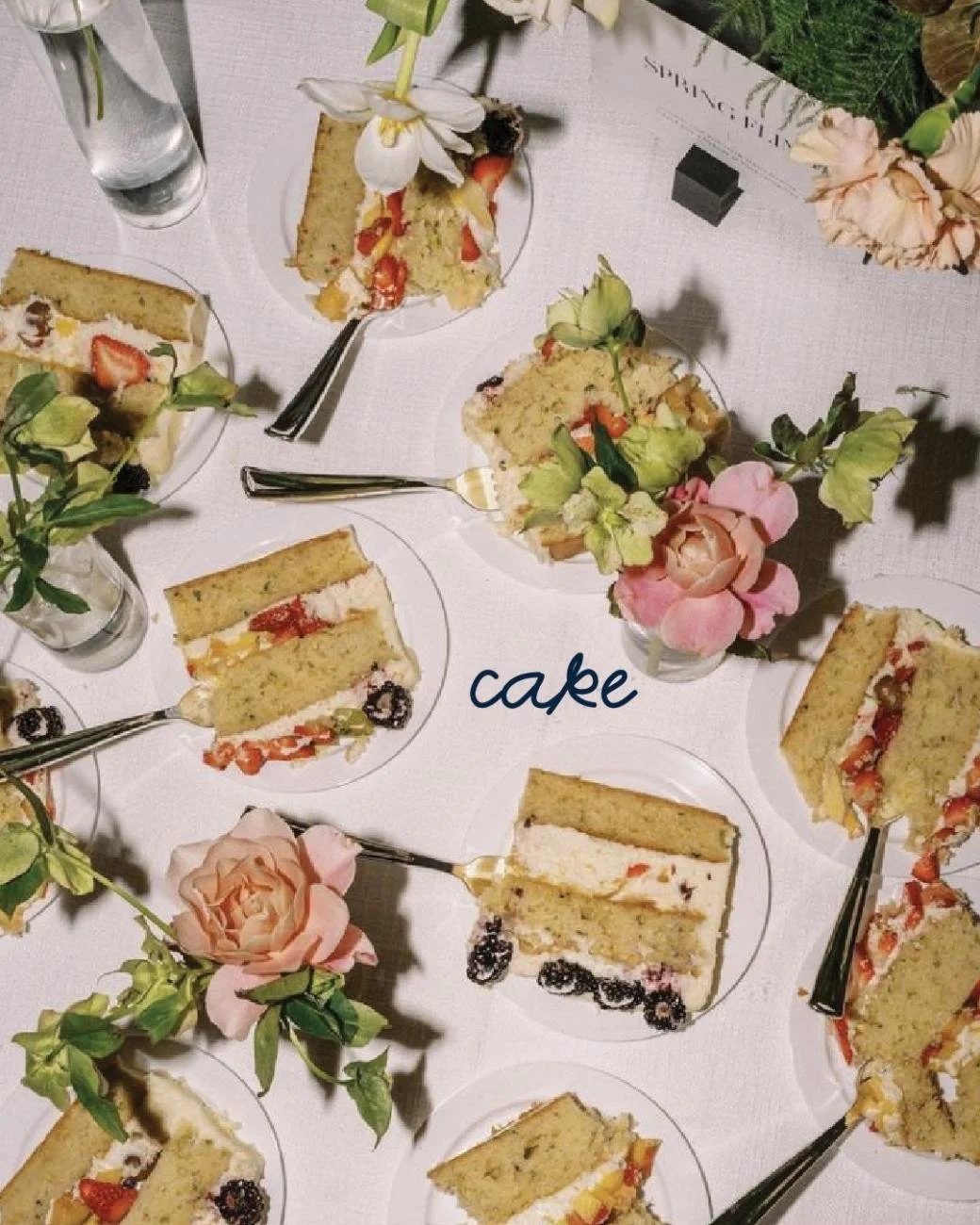 Slices of cake with cream, fruit, and berries on individual plates, arranged on a white tablecloth with floral decorations and a small sign that reads 'spring'.