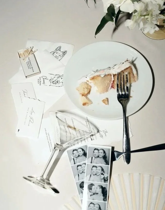 A used slice of cake on a white plate with a fork, a tipped-over empty wine glass, a photo booth strip with four black-and-white photos of a couple, wedding invitations, a small box with toothpicks, and a bouquet of white flowers on a white table.