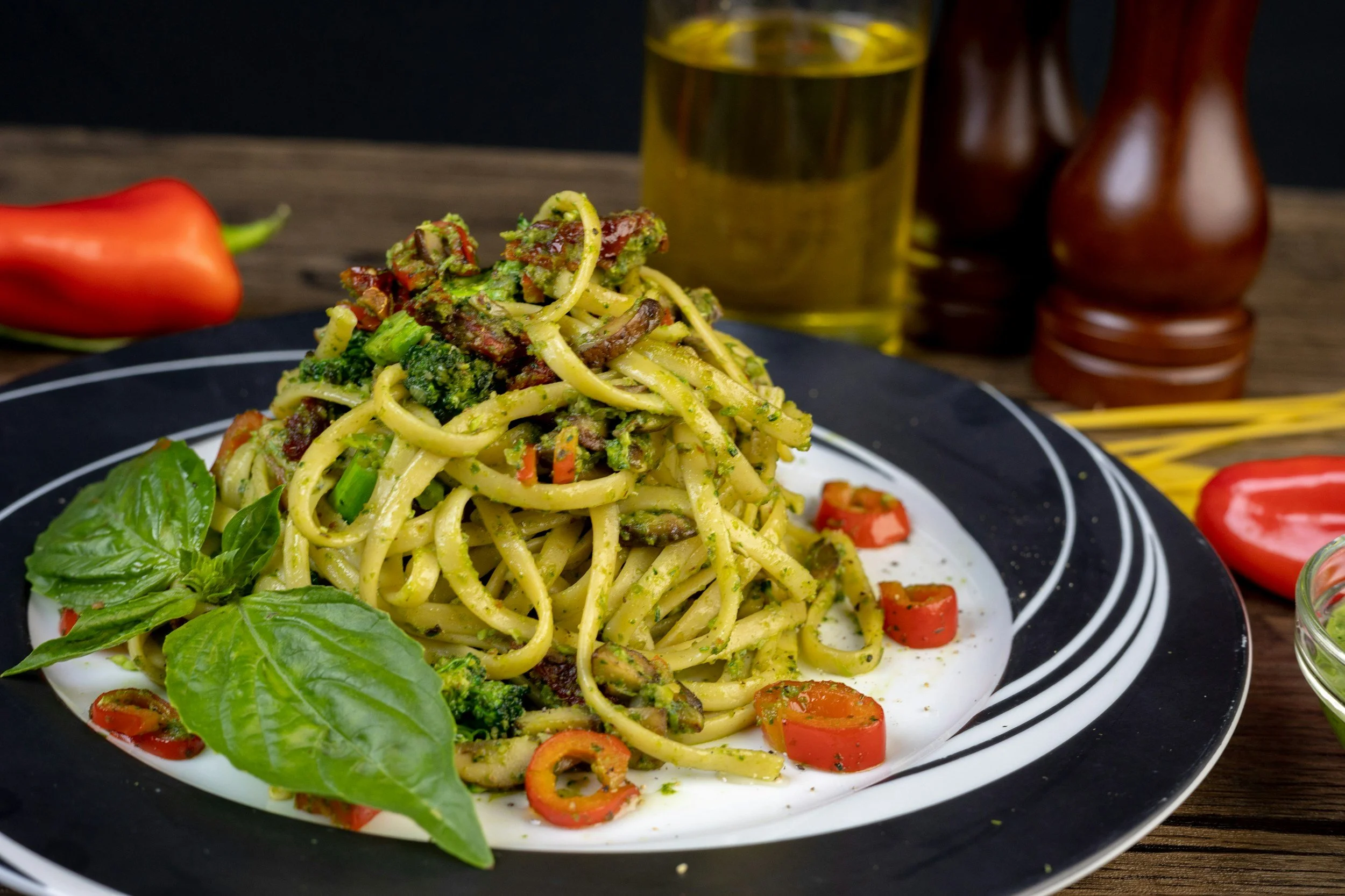 A plate of pasta with vegetables and basil leaves, garnished with cherry tomatoes and herbs, on a black and white plate on a wooden table with oil and pepper shakers in the background.