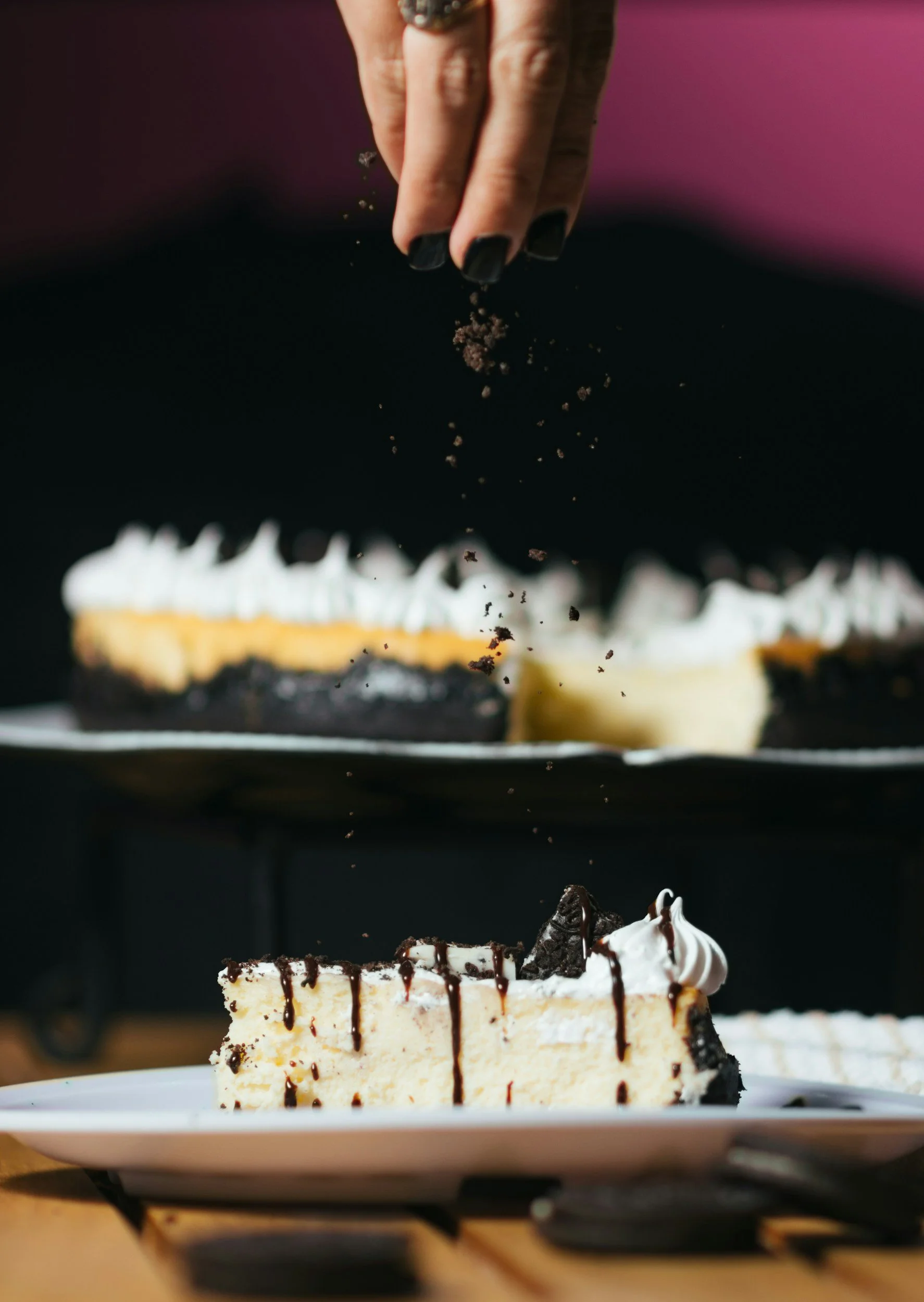 A slice of cheesecake with chocolate topping and whipped cream on a white plate, with a whole cake in the background, as someone sprinkles chocolate crumbs over the cake.