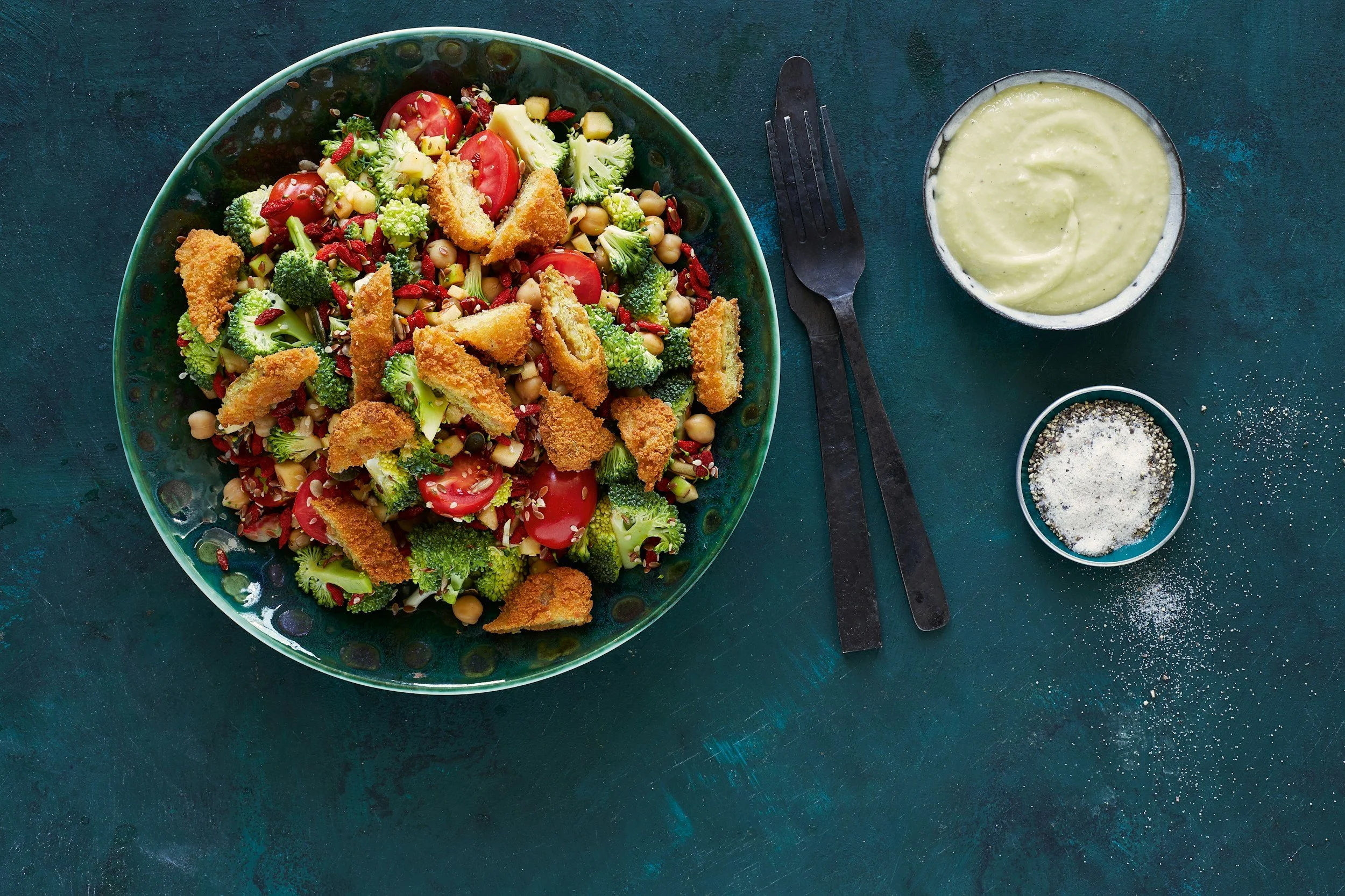 A large bowl of salad with broccoli, cherry tomatoes, chickpeas, corn, and breaded fried pieces, placed on a dark teal surface. To the right, there is a bowl of creamy dressing, a black-handled fork, and a black-handled knife.