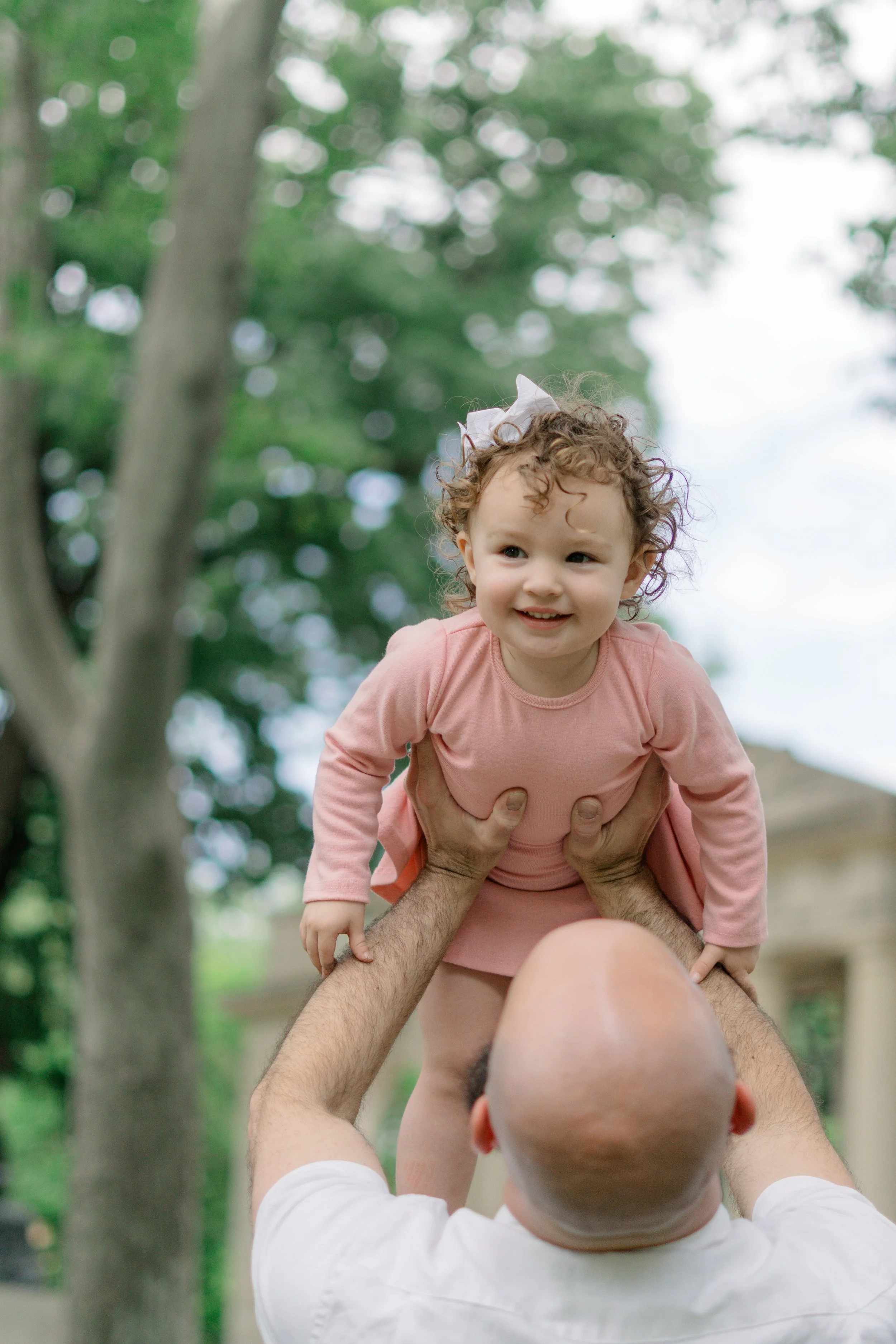 A man is lifting a young girl with curly hair and a pink bow, outdoors in a park with green trees in the background. The girl is smiling and looking to the side.
