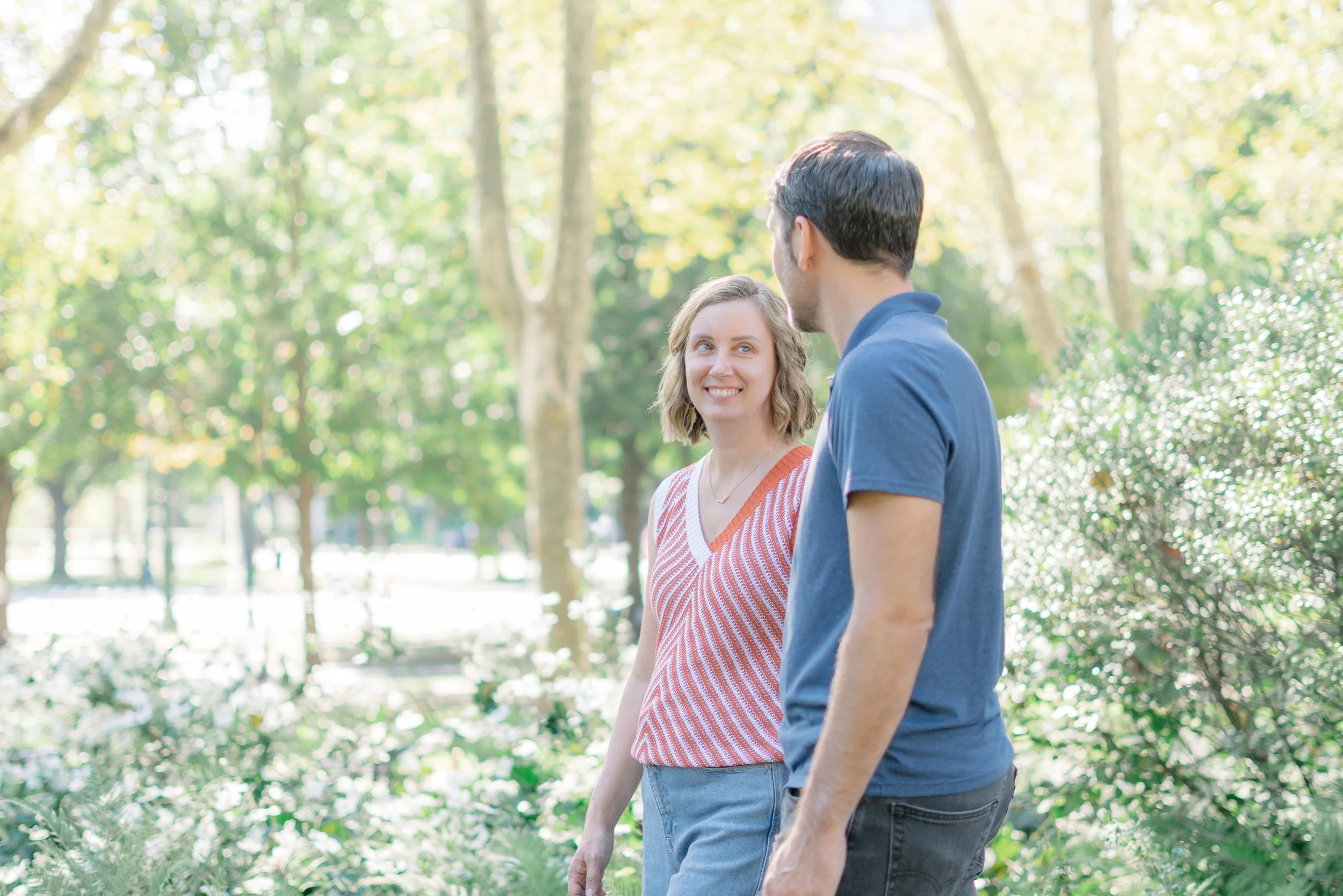 A young woman and a man are walking and talking in a park with trees and greenery, sunlight filtering through the leaves.