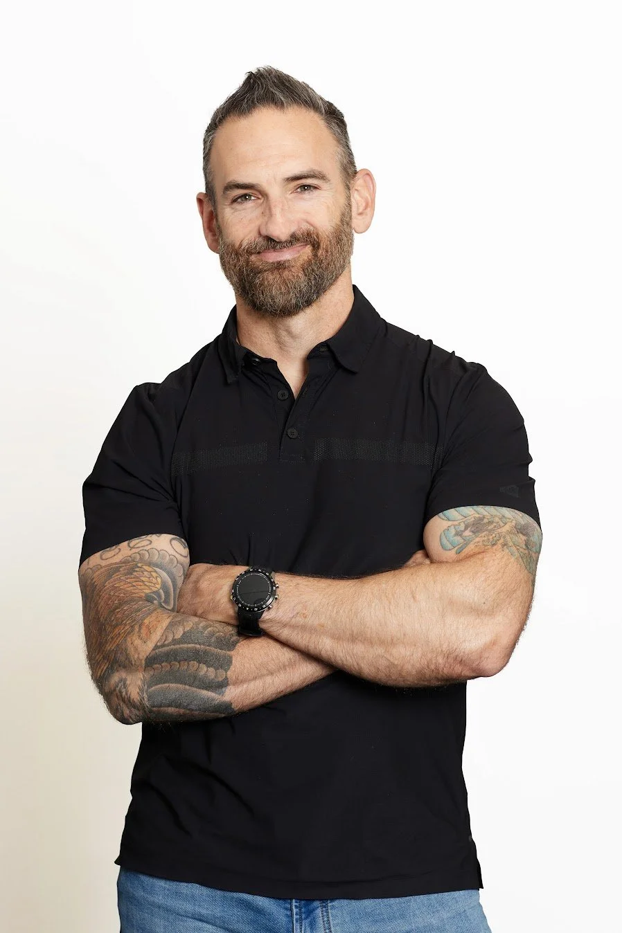 Man with a beard and tattoos on his arms, wearing a black polo shirt, standing with arms crossed and smiling against a white background.