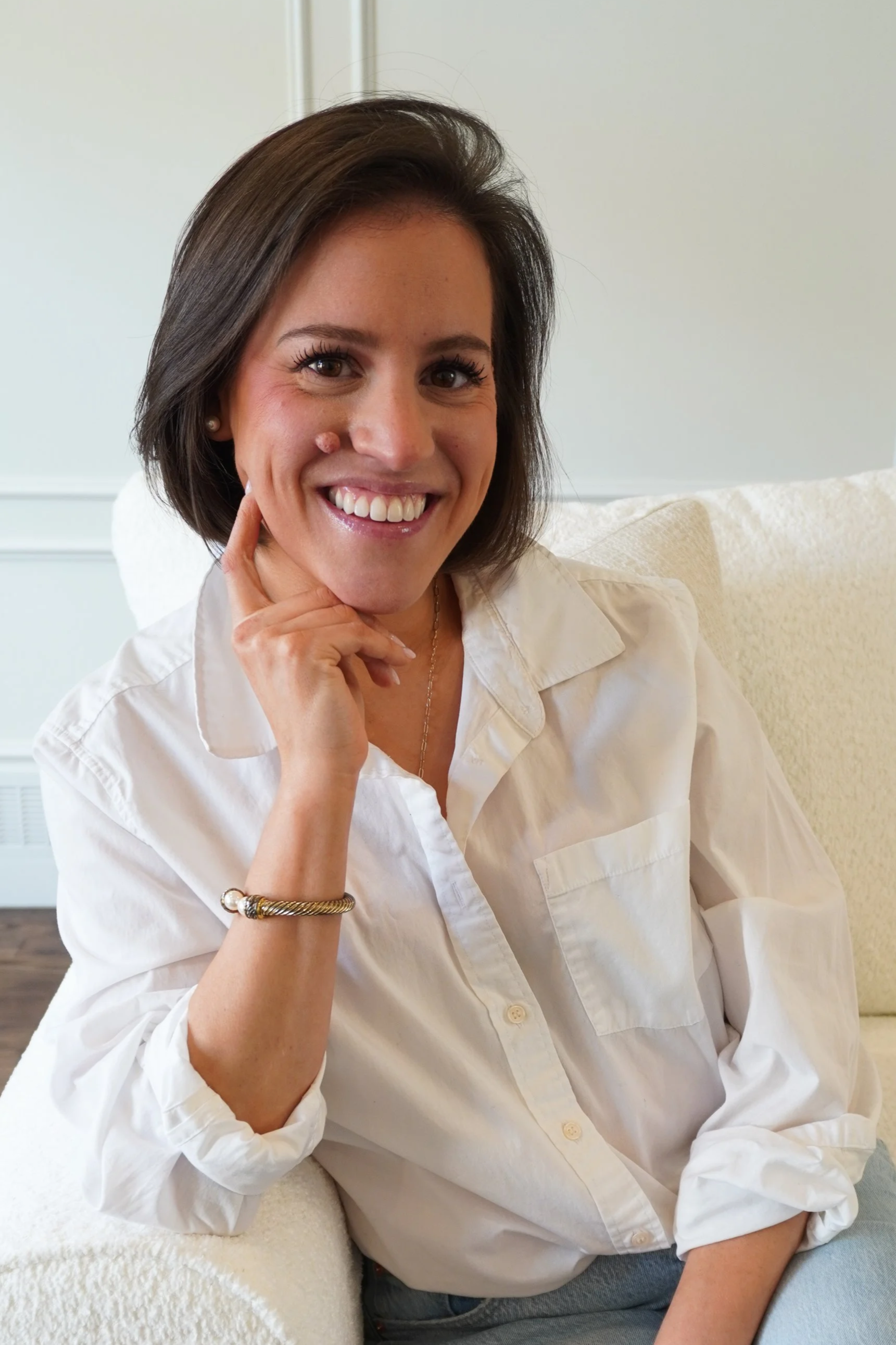 A woman with short dark hair smiling, resting her chin on her hand, sitting on a white sofa in a white shirt.