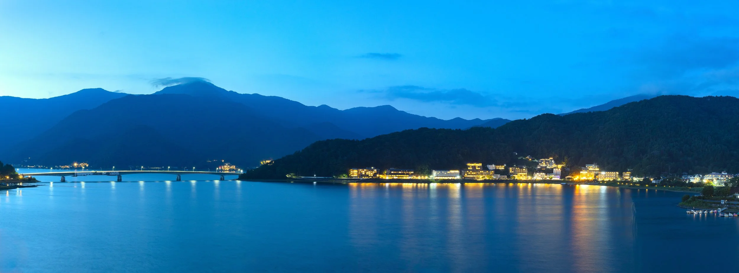 A night view of a river with city lights on the shore, mountains in the background, and a blue sky with a few clouds.