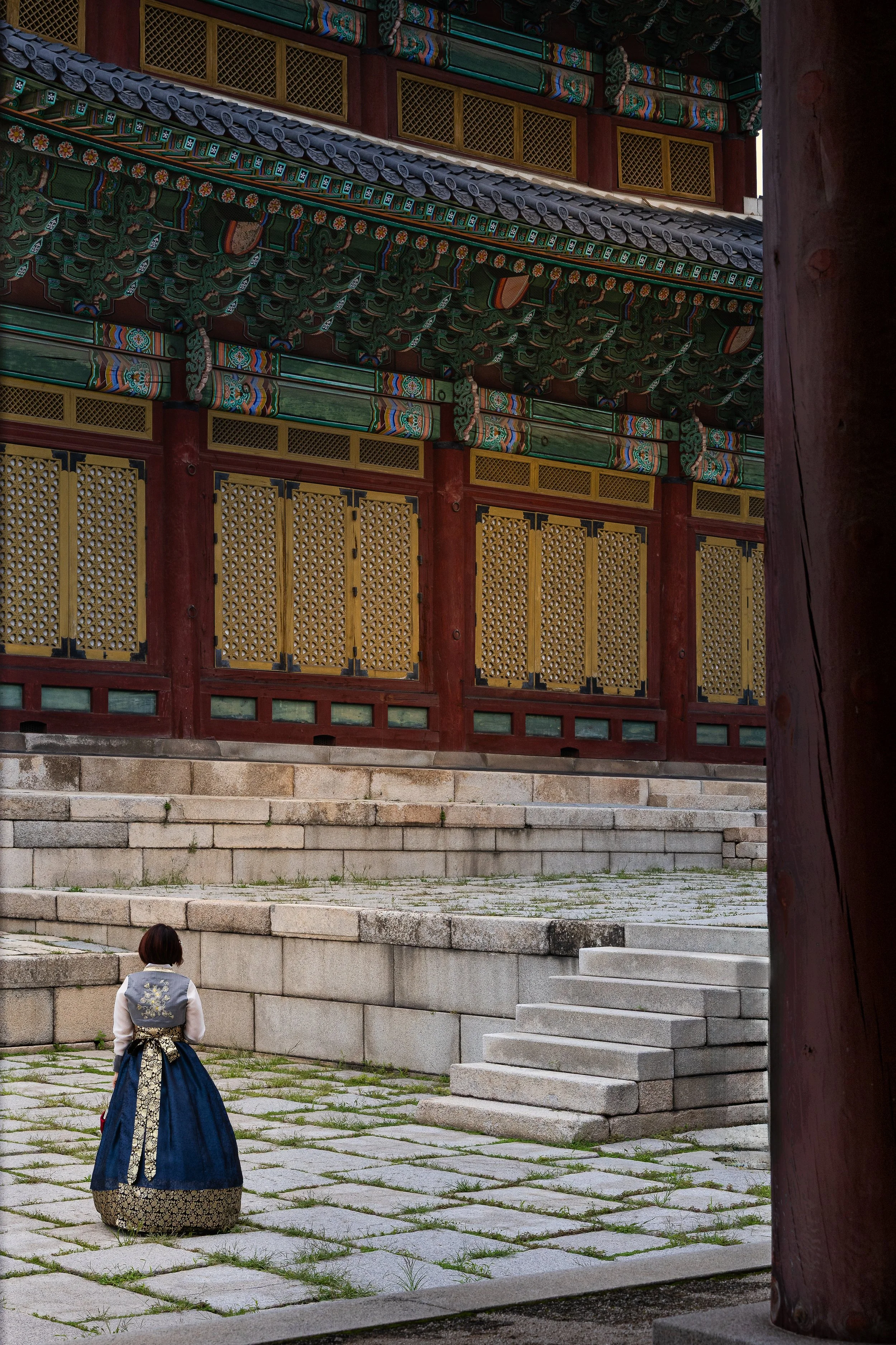 A person in traditional Korean clothing standing in front of a historic Korean building with intricate painted woodwork and stone steps.