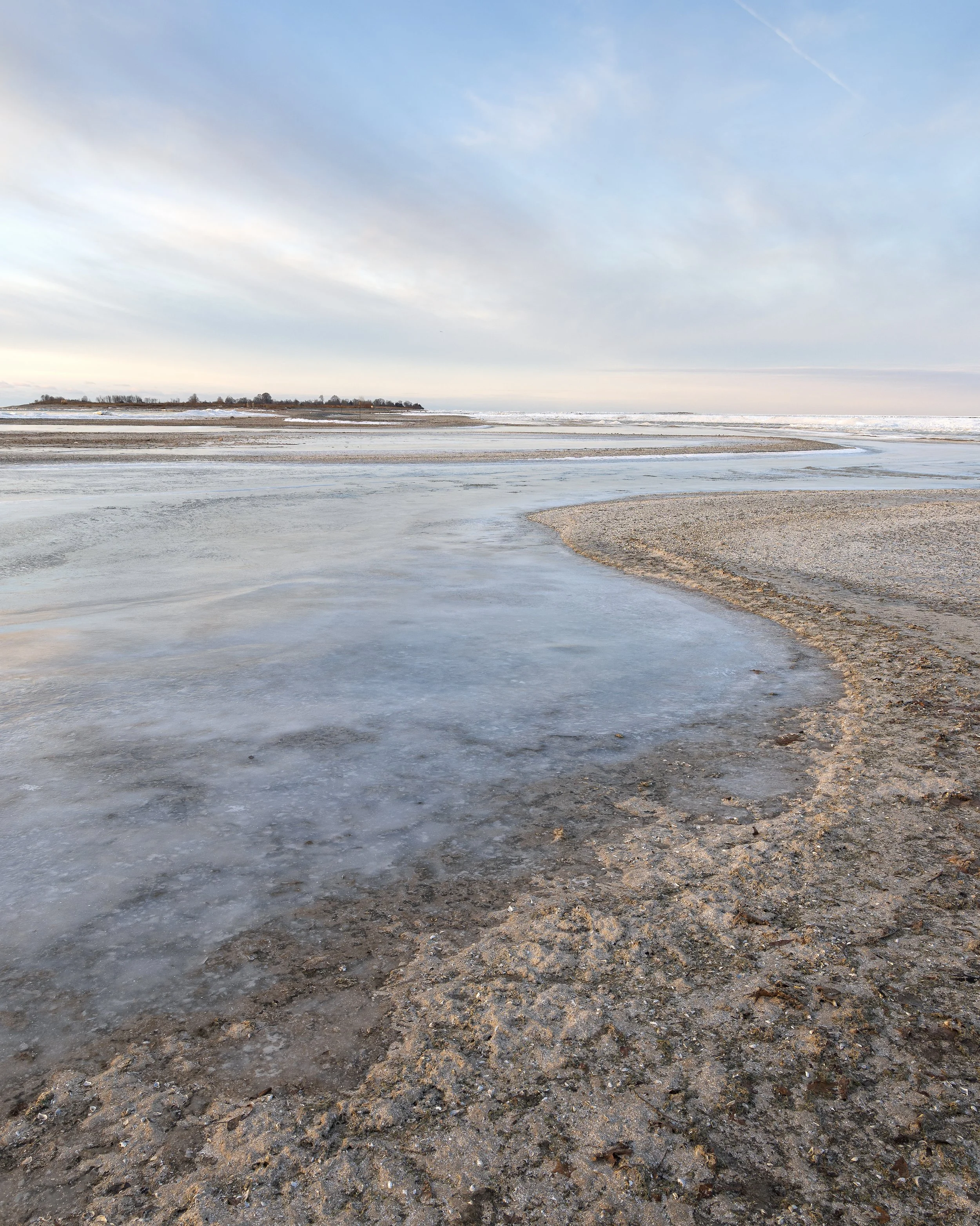 Cold shoreline with ice patches, sandy beach, and distant land under cloudy sky.