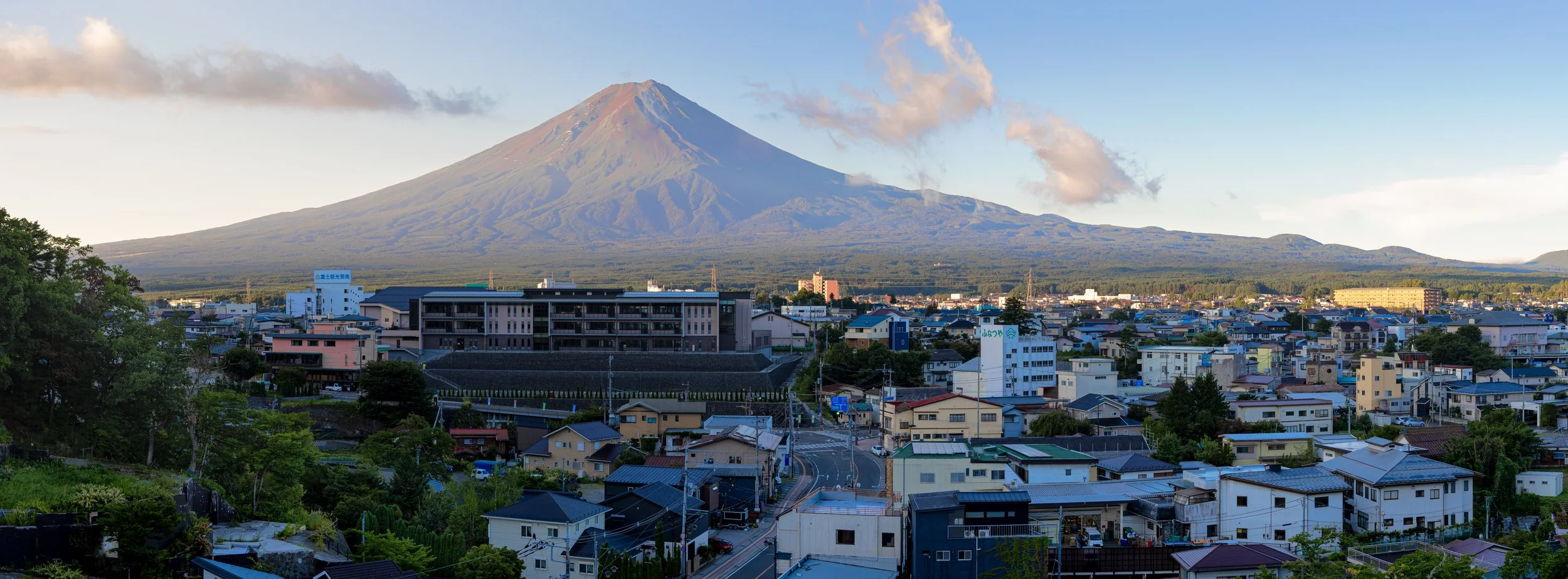 A scenic landscape of a town with Mount Fuji in the background, featuring a mix of houses, buildings, and greenery under a partly cloudy sky.