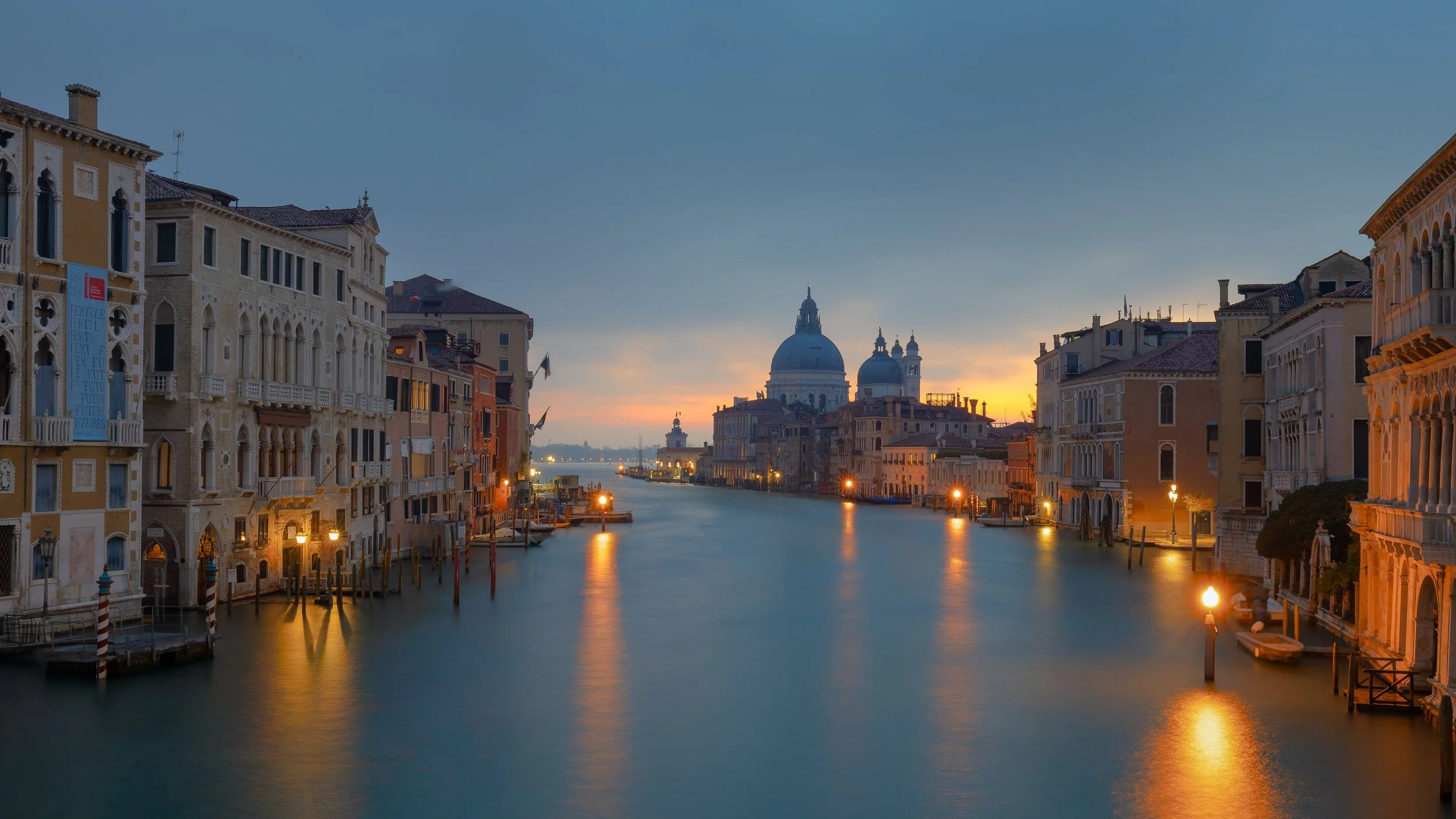 View of the Grand Canal in Venice, Italy, during sunset with historical buildings along the water and the domed Basilica di Santa Maria della Salute in the distance.