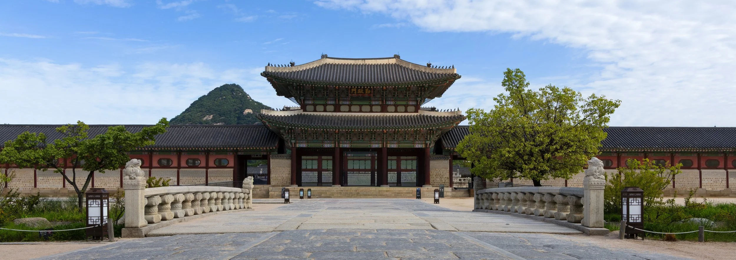 Traditional Korean palace gate with two-story tiled roof, stone lions, a stone bridge, and trees, with a mountain and blue sky in the background.